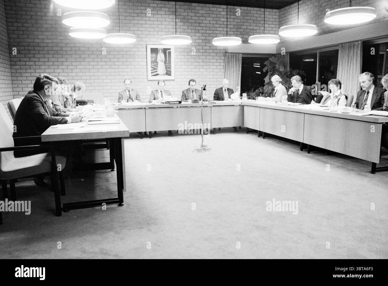 Chambre commune du Conseil. Réunion budgétaire de Bennebroek. Conseil municipal de Bennebroek Bennebroek, Whizgle News, Dutch Desk, pays-Bas, 1950 - 2000 on 21-11-1985. Ces rubriques apparaissent dans l'image. La scène capture une réunion formelle se déroulant autour d'une longue table rectangulaire qui domine le centre. Deux groupes distincts de personnes sont assis de chaque côté, tous habillés en tenue professionnelle, suggérant une atmosphère sérieuse et professionnelle. Les individus sont engagés dans la discussion, avec certains penchés en avant, stylos à la main, tandis que d'autres écoutent attentivement. L'éclairage est chaud et diffus, pr Banque D'Images