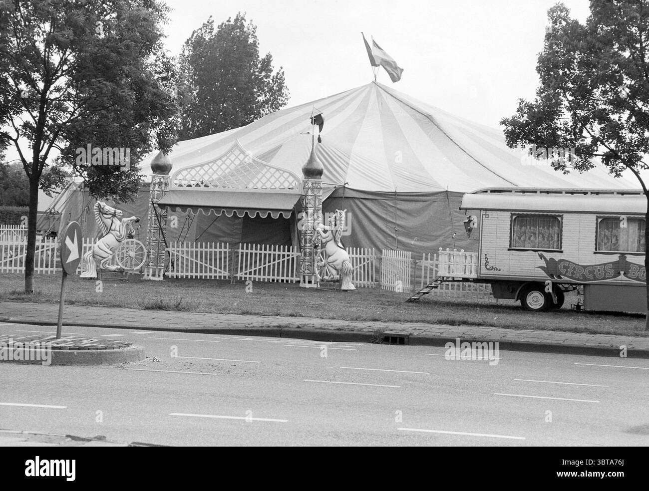 Circus Bongo Schalkwijk Haarlem pays-Bas, Whizgle News, Dutch Desk, pays-Bas, 1950 - 2000 le 10-06-1991. Voici les rubriques de l'image. La scène présente une grande tente de cirque, caractérisée par son auvent rayé audacieux dans des couleurs alternées de rouge et de blanc. La tente est bien en vue sur une tache ouverte d'herbe, entourée d'une clôture de piquet blanche basse qui ajoute une touche pittoresque au cadre. Flanquant la tente sont deux figures de clown fantaisistes, peintes de couleurs vives, prêtes dans des poses ludiques. Ils servent de décorations accrocheuses, rehaussant l'atmosphère festive. À l'avant Banque D'Images