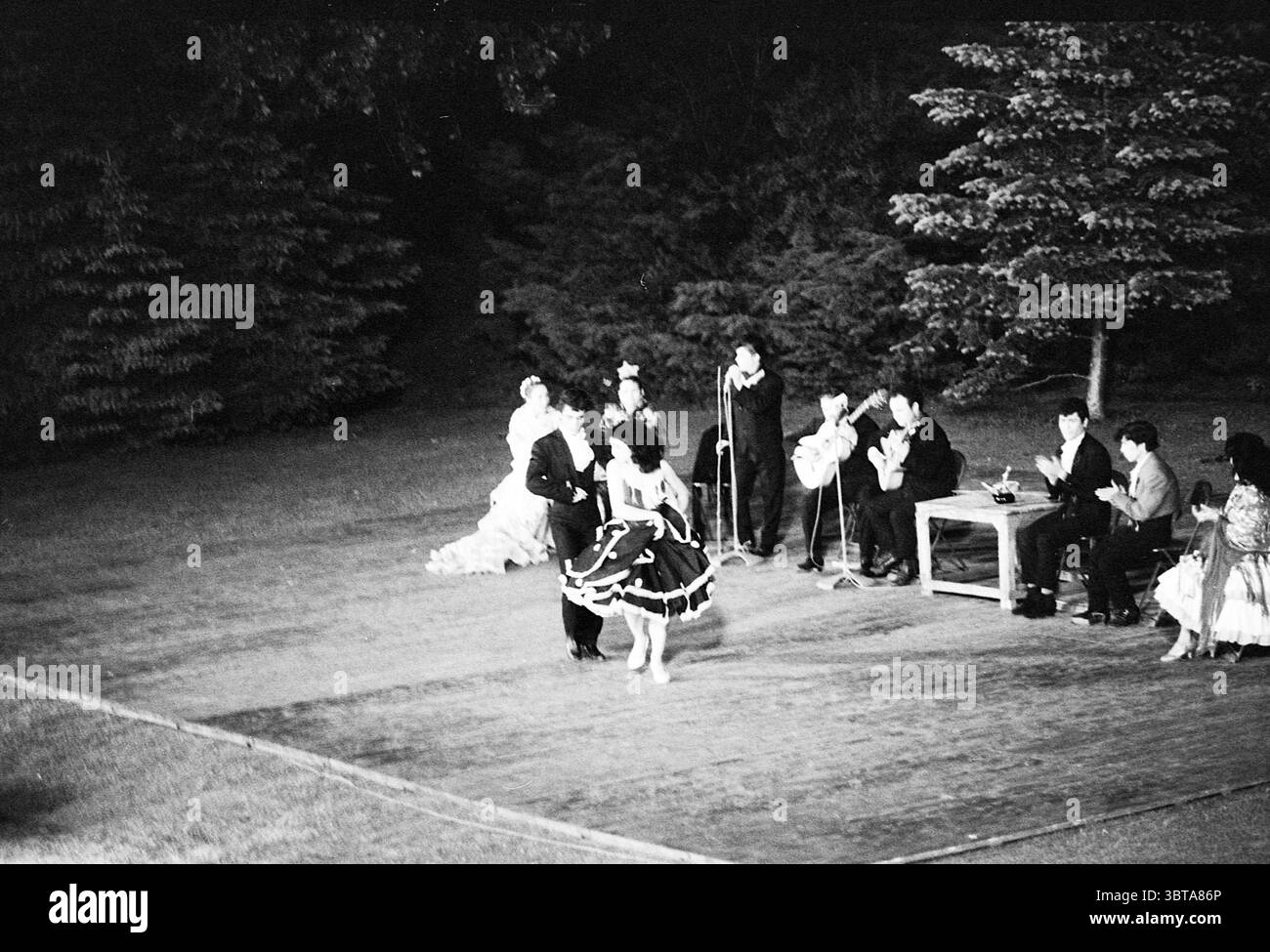 Danses espagnoles en plein air Bloemendaal danse Bloemendaal Hoge Duin en Daalseweg, Whizgle News, Dutch Desk, pays-Bas, 1950 - 2000 on 19-06-1962. L'image inclut ces rubriques. Dans un cadre extérieur légèrement éclairé, un groupe de personnes est réuni, créant une atmosphère de célébration ou de performance. La scène est ornée de parquet, jetant un charme rustique sur fond d'arbres assombrissants. Au premier plan, un couple danse énergiquement, la femme portant une robe noire vivante aux volants blancs qui accentuent ses mouvements. Son partenaire est habillé d'un costume noir formel Banque D'Images