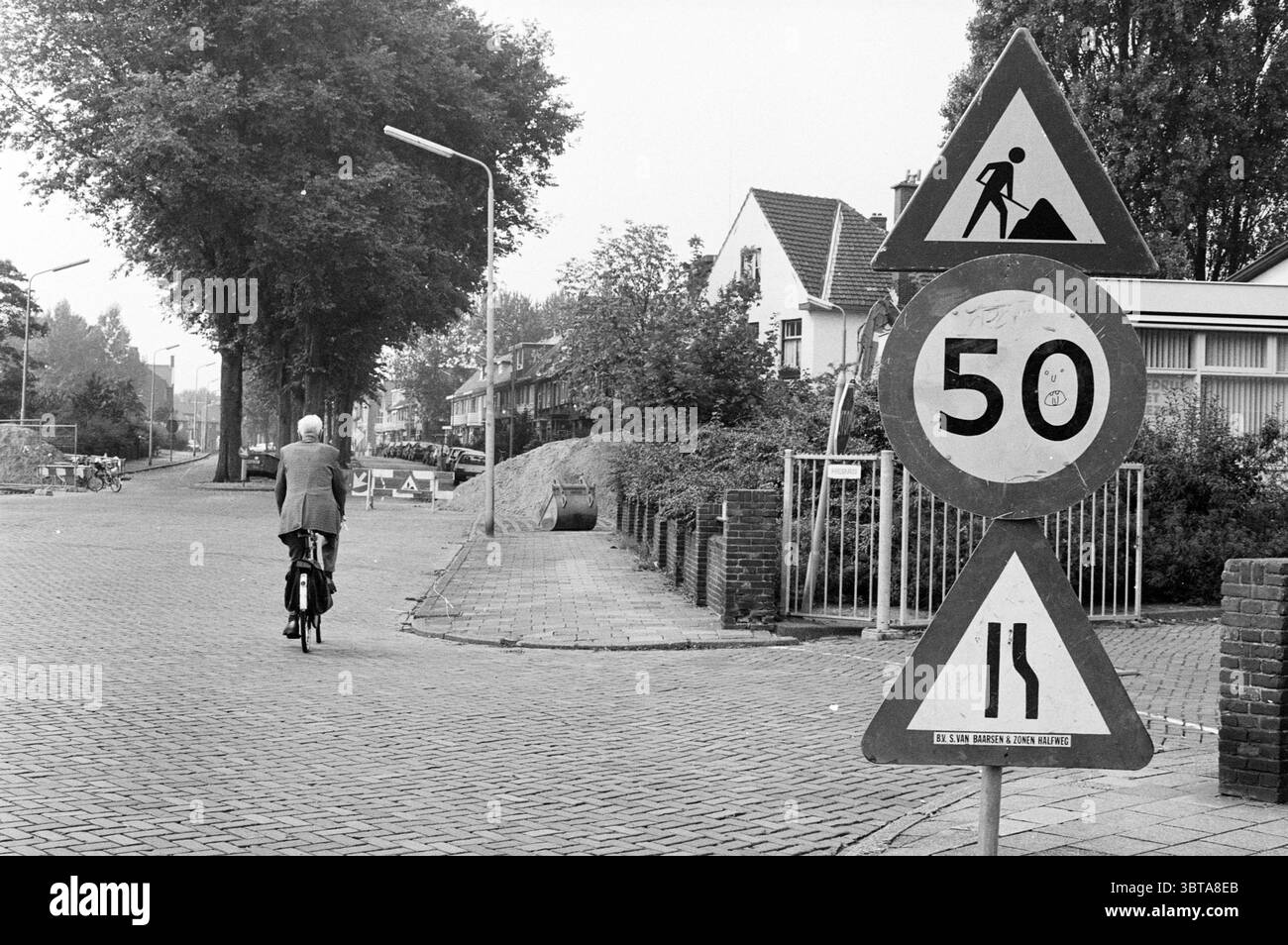 Panneau '50 km' Wijk aan Zeeërweg IJmuiden IJmuiden Wijk aan Zeeërweg pays-Bas, Whizgle News, Dutch Desk, pays-Bas, 1950 - 2000 le 07-09-1993. Ces rubriques sont illustrées dans l'image. La scène présente un cadre urbain tranquille, capturé dans une palette monochromatique qui souligne les contrastes entre la lumière et l'obscurité. Au premier plan, un cycliste âgé pédalera tranquillement sur un chemin pavé, montrant un sentiment de calme et de nostalgie. Habillé en tenue formelle, il semble incarner un mélange de tradition et de loisirs. À côté de lui, un panneau routier triangulaire est proéminent, marquant une zone de construction avec Banque D'Images