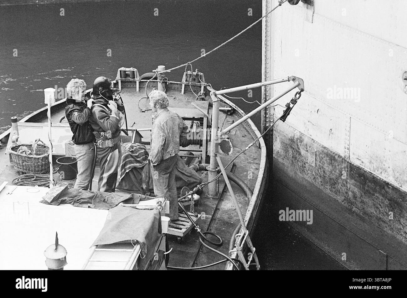 Travailler à l'écluse, Whizgle News, Dutch Desk, pays-Bas, 1950 - 2000 sur 13-10-1969. Ces rubriques apparaissent dans l'image. Dans un cadre maritime animé, trois personnes revêtent une tenue usée, révélatrice de leur environnement de travail pratique. La scène se déroule sur un bateau robuste, se balançant doucement sur des eaux calmes. La toile de fond présente un grand navire cargo imposant, sa surface métallique brillante légèrement sous la lumière du soleil silencieuse. Le premier personnage, vêtu d'un casque noir et d'un équipement pratique, dégage un air d'autorité et de concentration lorsqu'ils observent les opérations. A côté d'eux, un individu dans un cric en cuir Banque D'Images