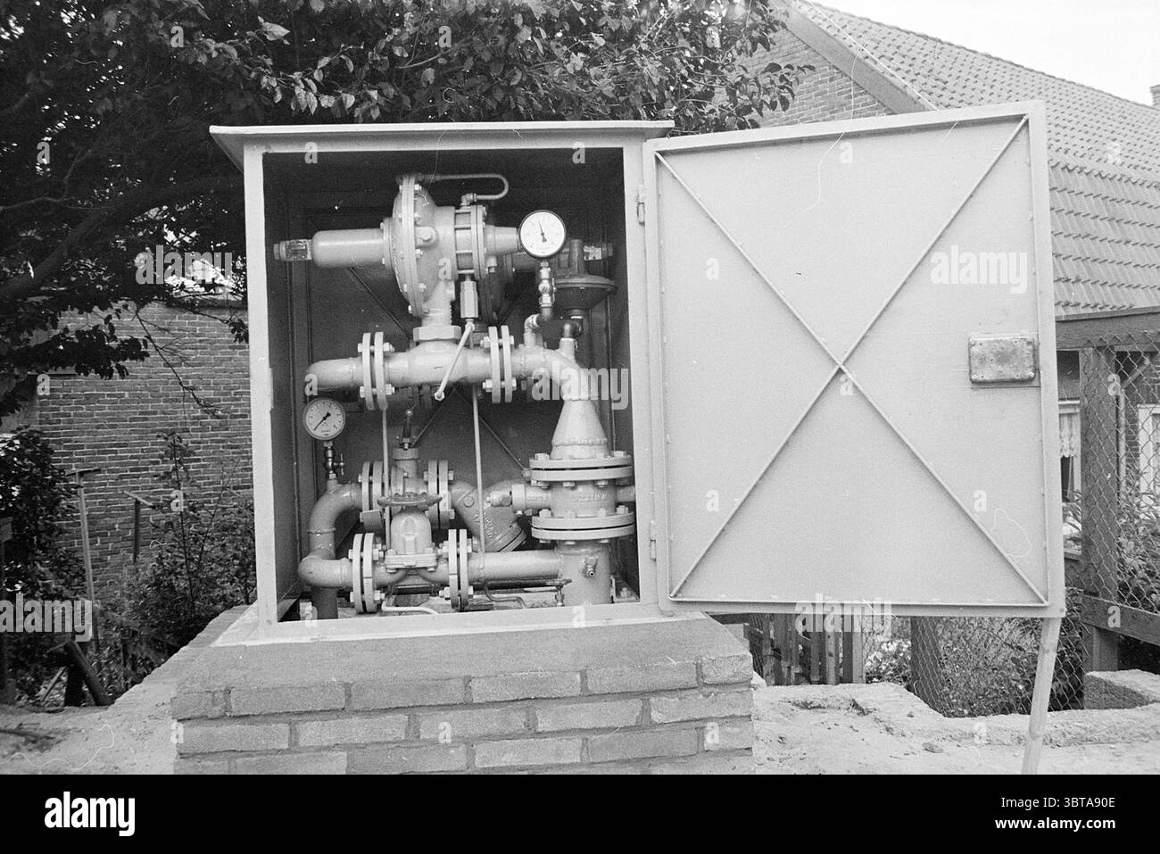 Armoire à gaz Spaarndam Gas, Whizgle News, Dutch Desk, pays-Bas, 1950 - 2000 le 02-08-1971. L'image inclut ces rubriques. La scène présente une boîte de service utilitaire positionnée en évidence sur un piédestal de brique, avec sa porte ouverte large pour révéler le fonctionnement intérieur. À l'intérieur de la boîte, un arrangement complexe de tuyaux métalliques et de vannes s'entrelacent, présentant un affichage mécanique. Divers jauges et cadrans, mesurant probablement la pression ou le débit, sont disposés stratégiquement, leurs faces en verre réfléchissant la lumière. La palette de couleurs est dominée par des nuances de gris atténuées, avec les composants métalliques apparaissant wor Banque D'Images
