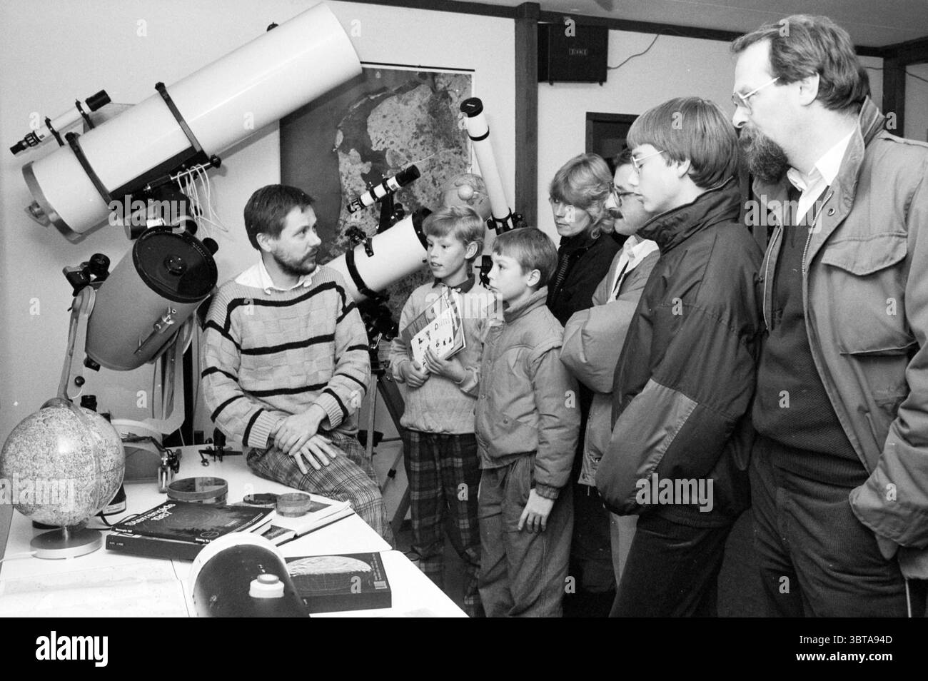 Journée portes ouvertes Observatoire Haarlem Observatoire Haarlem pays-Bas, Whizgle News, Dutch Desk, pays-Bas, 1950 - 2000 le 29-11-1987. Ce sont les éléments de l'image. La scène capture un groupe d'individus rassemblés autour d'une table présentant un assortiment d'équipements et de matériaux astronomiques. Au centre de la composition se trouve un homme assis à la table, faisant des gestes en expliquant quelque chose au public attentif. Il porte un pull rayé, ajoutant une touche décontractée mais engageante à son apparence. Devant lui se trouve un globe, qui représente probablement des corps célestes ou la Terre elle-même, Alo Banque D'Images