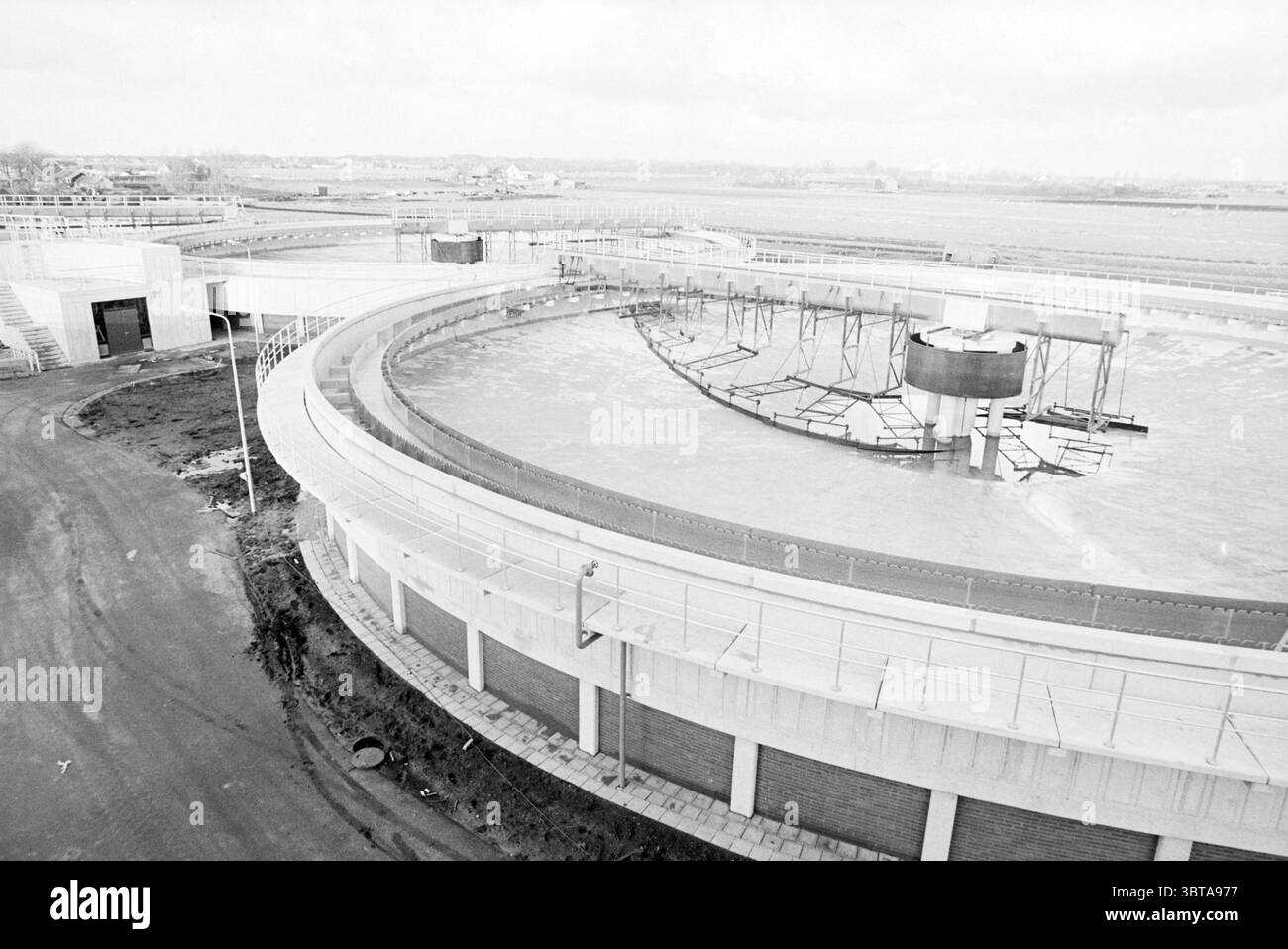 Station de traitement de l'eau Zwaanshoek Water Zwaanshoek, Whizgle News, Dutch Desk, pays-Bas, 1950 - 2000 on 16-12-1980. L'image contient ces rubriques. La scène présente une grande installation industrielle, caractérisée par ses structures circulaires et linéaires. Les éléments dominants sont de grands réservoirs circulaires, probablement une partie d'une station d'épuration des eaux usées, avec des passerelles circulaires et des chemins d'accès rayonnant vers l'extérieur. Ces réservoirs sont remplis d'eau, réfléchissant la lumière ambiante et créant une surface chatoyante. Le cadre est vaste, avec une toile de fond d'un ciel nuageux qui projette une lumière douce et diffuse o Banque D'Images