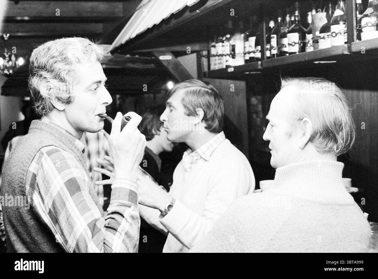 Diverses personnes en conversation, Whizgle News, Dutch Desk, pays-Bas, 1950 - 2000 le 07-05-1979. L'image montre ces rubriques. Dans un intérieur chaleureusement éclairé, un rassemblement confortable se déploie dans un cadre rustique. L’espace présente des poutres en bois et des étagères bordées de bouteilles, évoquant un sentiment de confort et de convivialité. Au premier plan, un homme se tient à gauche, le visage encadré de cheveux clairs ondulés, vêtu d'un pull à motifs. Il tient un tuyau, apparemment perdu dans la pensée ou la conversation, exsudant un comportement détendu. Le sourire subtil sur son visage fait allusion à un moment léger. Banque D'Images