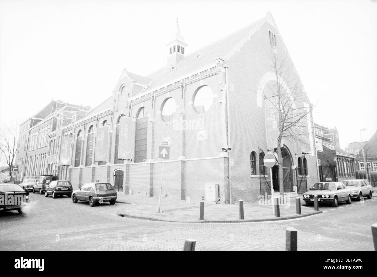 Église extérieure libre. Church [Église libre réformée de Vestekerk], Whizgle News, Dutch Desk, pays-Bas, 1950 - 2000 le 24-01-1996. Voici les rubriques de l'image. La scène représente un bâtiment historique caractérisé par son style architectural distinctif. La structure possède une façade en briques rouges avec des détails complexes, y compris des fenêtres cintrées qui présentent des motifs circulaires et un toit à pignon proéminent, couronné par une petite flèche. La palette de couleurs globale est en niveaux de gris, créant une atmosphère intemporelle et légèrement nostalgique. Entourant le bâtiment, une rue est bordée de véhicules stationnés, showcas Banque D'Images