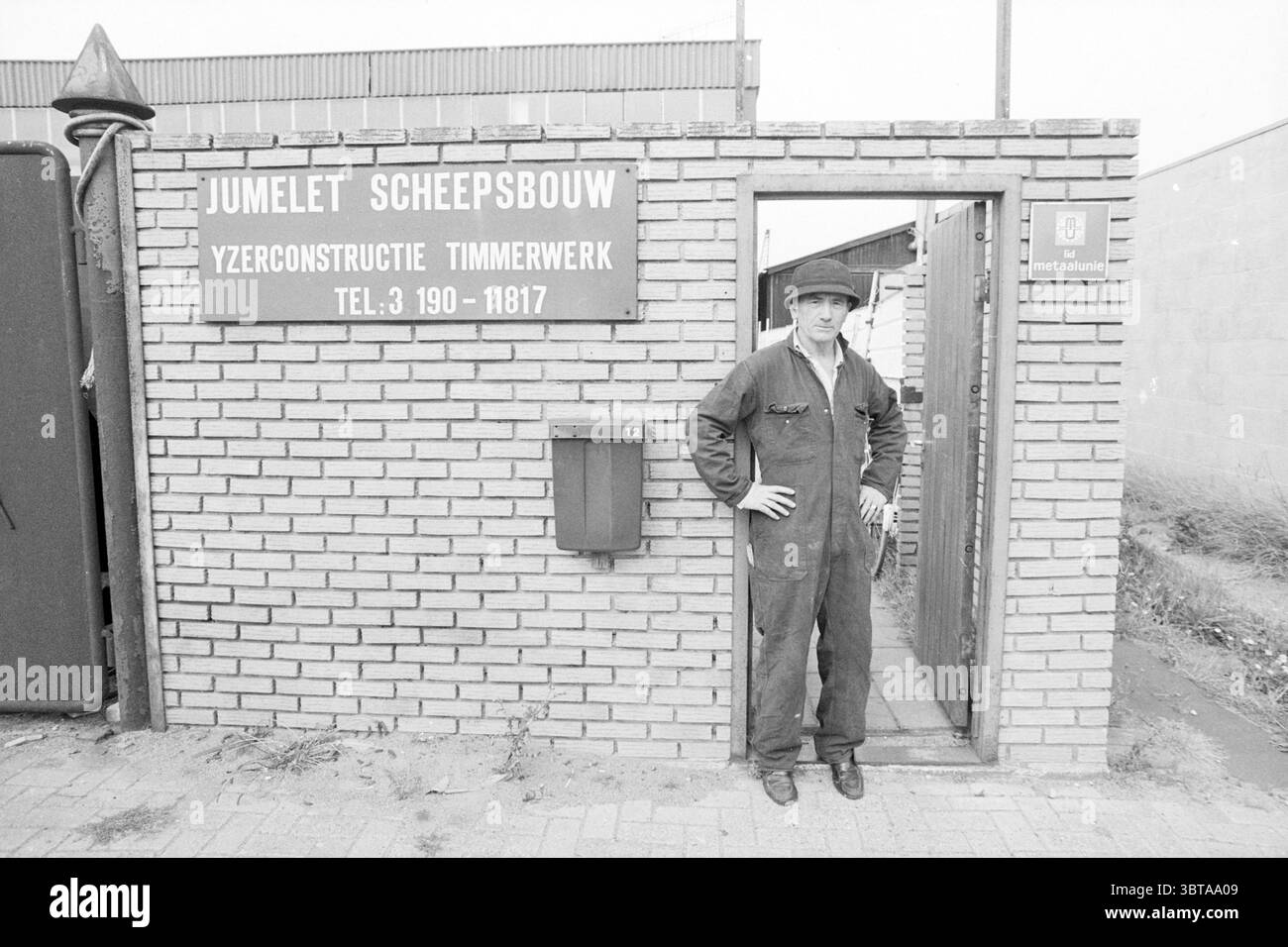 M. Jumelet Shipbuilder IJmuiden Shipbuilding shipping IJmuiden pays-Bas, Whizgle News, Dutch Desk, pays-Bas, 1950 - 2000 on 22-08-1988. L'image montre ces rubriques. La scène présente un cadre industriel, caractérisé par un mur de briques altérées qui sert de toile de fond. Une porte, légèrement entrouverte, mène à un espace de travail modeste ou à une installation. Les briques sont une teinte sourde, faisant allusion à l'âge et à l'usure, avec certaines zones montrant des signes de décoloration et de décollement de la peinture. Debout dans la porte est un individu vêtu d'un uniforme de travail sombre, qui semble pratique et robuste, adapté au manua Banque D'Images