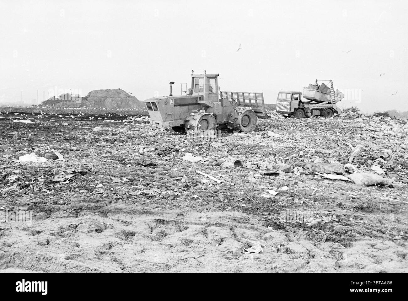 Aperçu de Spaarnwoude déchets de décharge Spaarnwoude, Whizgle News, Dutch Desk, pays-Bas, 1950 - 2000 sur 01-12-1980. L'image inclut ces rubriques. La scène présente un paysage désolant dominé par une grande étendue de déchets. Deux véhicules puissants se dressent au milieu des débris : un chargeur frontal, robuste et imposant, a son godet légèrement relevé, suggérant qu'il collecte les ordures. Sa couleur jaune sourde contraste avec le brun grisâtre du sol. Le deuxième véhicule, plus petit et situé plus en arrière, semble être un camion à benne basculante ; sa conception indique qu’il est construit pour des tâches lourdes. Th Banque D'Images