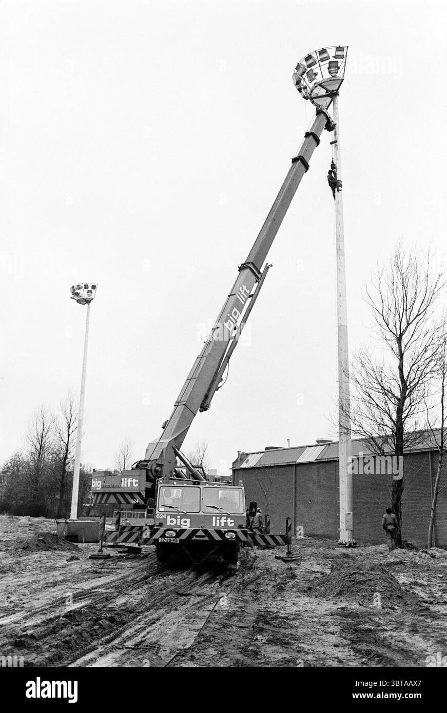 Installation de lampadaires au Pim Mulier Stadium Sports Fields Lighting Haarlem pays-Bas, Whizgle News, Dutch Desk, pays-Bas, 1950 - 2000 on 07-02-1980. Ces rubriques apparaissent dans l'image. La scène représente un chantier de construction caractérisé par une atmosphère industrielle dure. Une grande grue domine le premier plan, son long bras s'étendant vers le haut en direction d'un poteau haut et élancé orné d'un faisceau circulaire de projecteurs. Le corps de la grue révèle un design géométrique audacieux dans un schéma de couleurs contrastées, mettant probablement l'accent sur l'utilité et la force. En arrière-plan, on peut voir la silhouette ombragée Banque D'Images