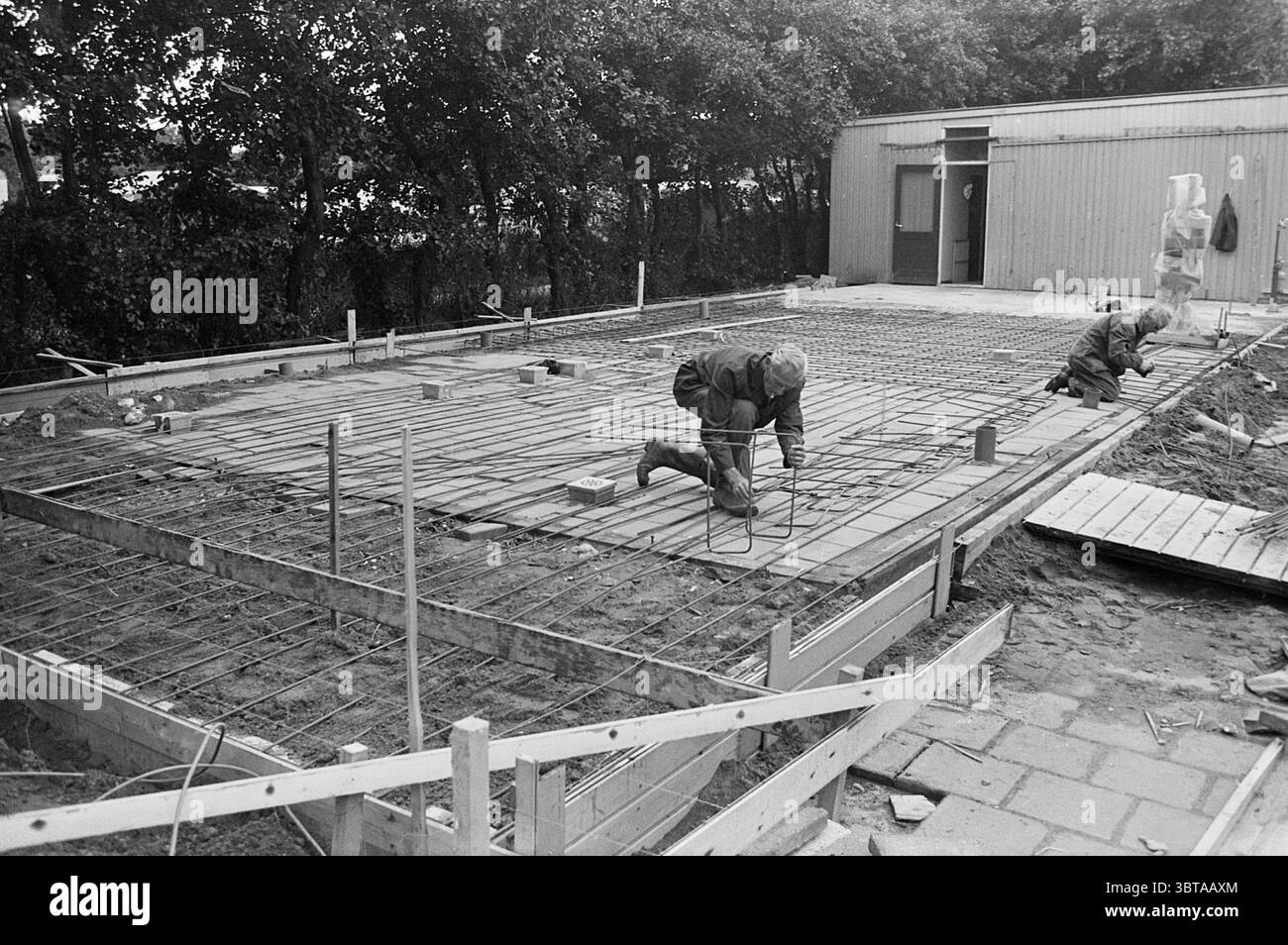 Construction du vestiaire Jong Hercules Beverwijk. Construction construction Pits, Whizgle News, Dutch Desk, pays-Bas, 1950 - 2000 sur 31-07-1974. Voici les rubriques de l'image. La scène représente un chantier de construction, où deux ouvriers sont engagés dans la pose des bases pour une structure. Le premier plan est dominé par un cadre de poutres en bois et de barres d'armature en métal, indiquant que la fondation est en cours de construction. Les travailleurs, habillés de vêtements sombres et pratiques, se concentrent intensément sur leurs tâches, l'un accroupissant et manipulant les matériaux tandis que l'autre se tient debout, ajustant le Re Banque D'Images