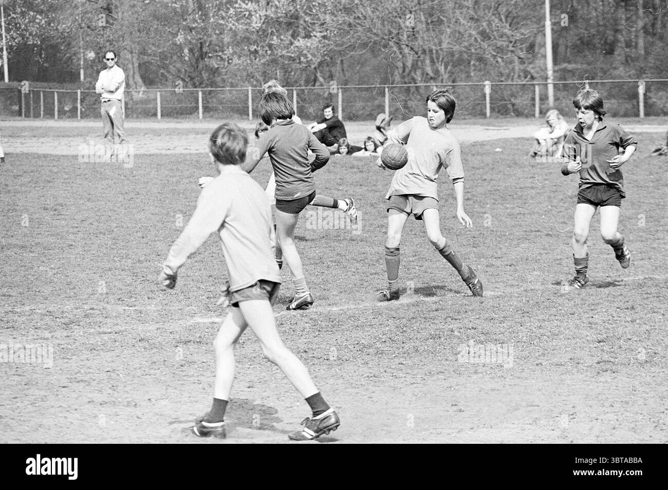 Football scolaire dans IJmuiden Football scolaire, Whizgle News, Dutch Desk, pays-Bas, 1950 - 2000 le 10-04-1974. Ces rubriques apparaissent dans l'image. La scène capture un moment animé sur un terrain herbeux où un groupe d'enfants sont engagés dans une partie de football. Le cadre est une zone ouverte avec de l'herbe fraîchement coupée, parsemée de taches de terre usée où les joueurs ont couru. Au premier plan, deux garçons sont en évidence. L'un est en mouvement, à mi-foulée, alors qu'il dribble le ballon de football avec détermination concentrée, portant un maillot gris ample et un short long genou. Ses chaussettes sont remontées Banque D'Images