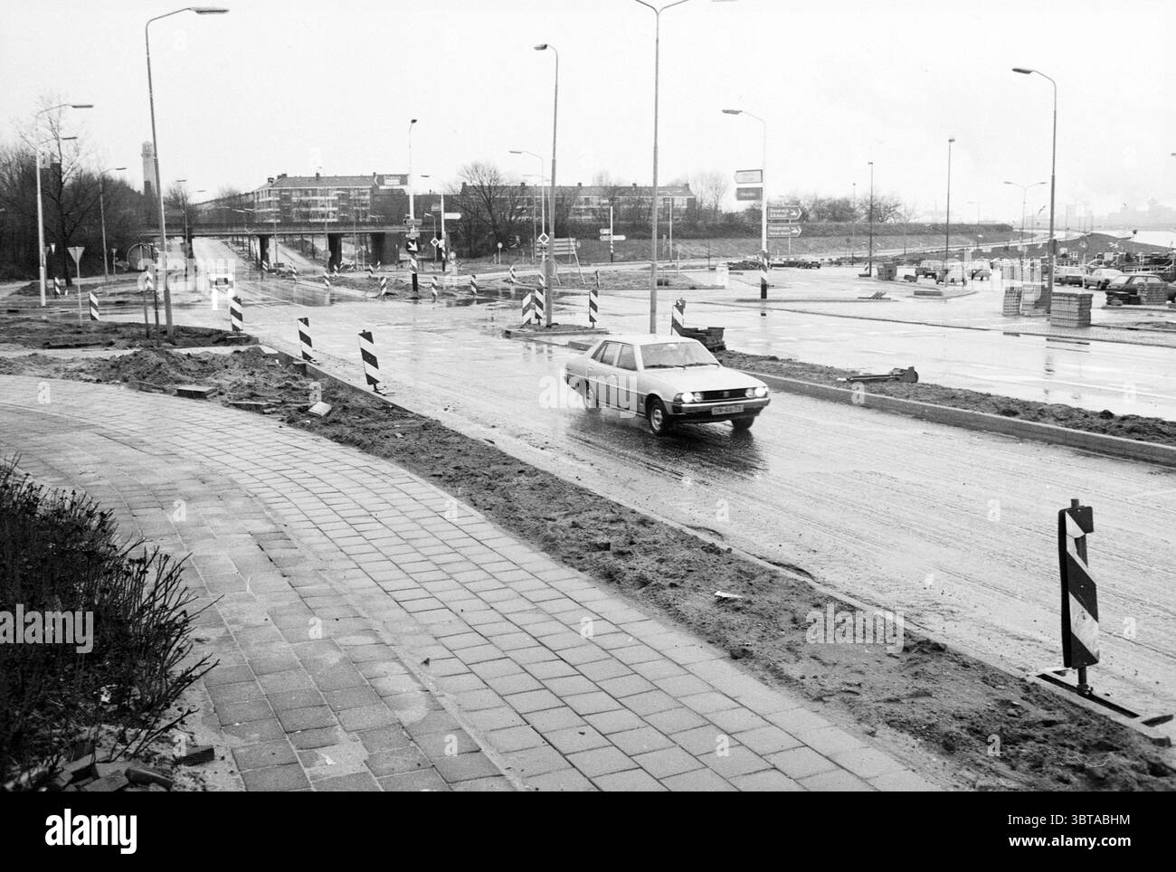 Aperçu Pontplein IJmuiden Pleinen Velsen-Zuid Pontplein, Whizgle News, Dutch Desk, The Netherlands, 1950 - 2000 sur 21-12-1986. L'image contient ces rubriques. La scène présente un jour gris et couvert, où l'humidité de la pluie récente est palpable. La route est un mélange d'asphalte humide et de sections légèrement boueuses, reflétant le ciel sombre au-dessus. D'un côté, un trottoir soigneusement pavé court parallèlement à la chaussée, bordé par des parcelles d'herbe clairsemées et de petits arbustes qui semblent lutter dans l'environnement froid. Une voiture seule roule le long de la route boueuse, sa carrosserie reflétant les tons discrets du Banque D'Images