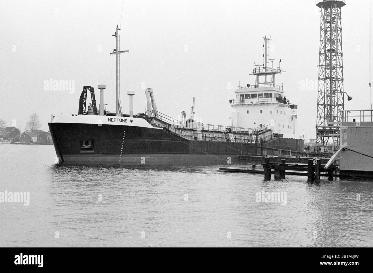 Neptune : drague à trémie fendue à Scheepswerf Stapel Spaarndam Schepen Spaarndam, Whizgle News, Dutch Desk, pays-Bas, 1950 - 2000 on 07-04-1983. Ce sont les éléments de l'image. La scène présente un grand cargo, bien visible comme sujet central. Sa coque est peinte d'un noir dur, contrastant avec le gris discret d'une atmosphère brumeuse. Le pont supérieur comporte une superstructure blanche ornée d'une série d'antennes et d'équipements, indiquant sa fonctionnalité pour les opérations maritimes. Autour du navire se trouve une étendue d'eau calme, reflétant la lumière douce et diffuse Banque D'Images