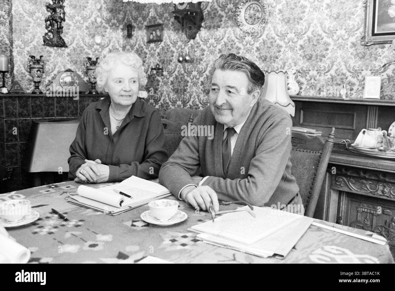 M. Braam auteur d'un livre à IJmuiden avec sa femme à la table dans le salon IJmuiden pays-Bas, Whizgle News, Dutch Desk, pays-Bas, 1950 - 2000 le 08-02-1985. Voici les rubriques de l'image. Dans une pièce chaleureusement décorée, deux personnes s'assoient à une table ornée d'une nappe à motifs floraux. Les couleurs de la nappe sont douces, donnant une douce chaleur au décor. La table est sertie de tasses délicates, suggérant un moment de thé ou de café, ajoutant à l'atmosphère accueillante. D'un côté, une femme âgée avec les cheveux blancs bouclés et un comportement gentil porte un Banque D'Images