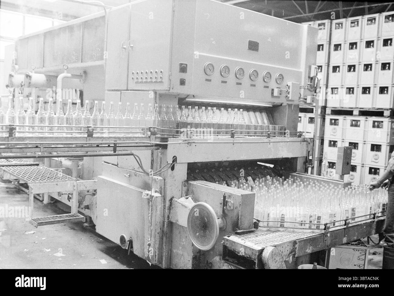 Lave-bouteilles Loots Lemonade Drinks and Liquor Stores, Whizgle News, Dutch Desk, pays-Bas, 1950 - 2000 le 21-06-1982. Voici les rubriques de l'image. La scène représente un environnement industriel très fréquenté, en particulier une installation de mise en bouteille ou d'emballage. Au premier plan, une grande machine domine l’espace, son corps métallique réfléchissant la lumière ambiante. La machine est équipée de divers cadrans et boutons, suggérant un niveau de complexité et de précision dans son fonctionnement. À gauche, une série de bouteilles en verre transparent sont alignées, debout lorsqu'elles se déplacent le long d'un tapis roulant. Le bott Banque D'Images