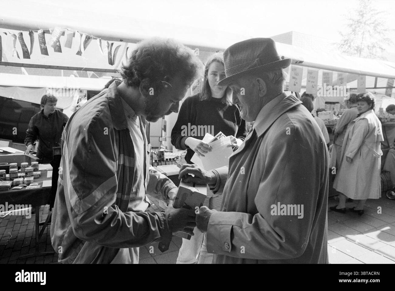 Collection pour Cameroun Marktplein Hoofddorp Collection Hoofddorp Marktplein pays-Bas, Whizgle News, Dutch Desk, pays-Bas, 1950 - 2000 on 04-09-1986. Voici les rubriques de l'image. Dans une scène de marché dynamique, un mélange de personnes s'engagent dans des interactions animées au milieu d'étals regorgeant de produits divers. L'atmosphère est animée mais commune, suggérant un point de rassemblement local. Au premier plan, deux hommes sont engagés dans une transaction. L'un, avec une coiffure bouleversée et portant une veste décontractée, se penche intentionnellement en avant, examinant une certaine monnaie. L'autre, un vieux doux Banque D'Images