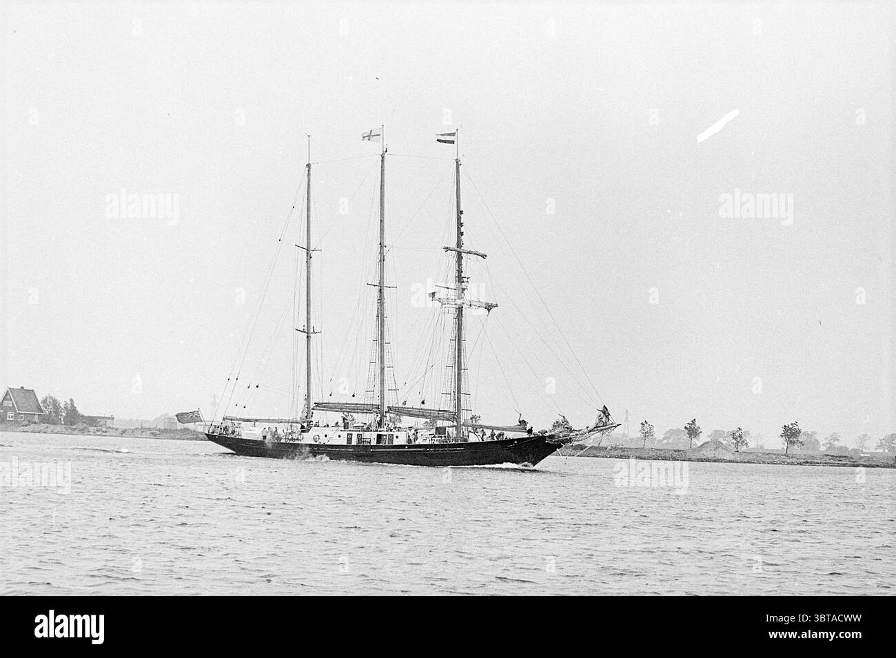Goélette IJmuiden Ships, Whizgle News, Dutch Desk, pays-Bas, 1950 - 2000 le 17-07-1972. L'image montre ces rubriques. La scène représente un majestueux voilier glissant sur des eaux calmes. Le navire comporte trois mâts hauts et complexes ornés de voiles, dont certaines sont déployées, attrapant la douce brise. La coque du navire est peinte dans une couleur profonde et sombre qui contraste frappamment avec les teintes plus claires de l'eau en contrebas. En arrière-plan, de faibles silhouettes d'arbres et peut-être de bâtiments bordent le rivage, suggérant un cadre côtier tranquille. Le paysage au-delà apparaît Banque D'Images