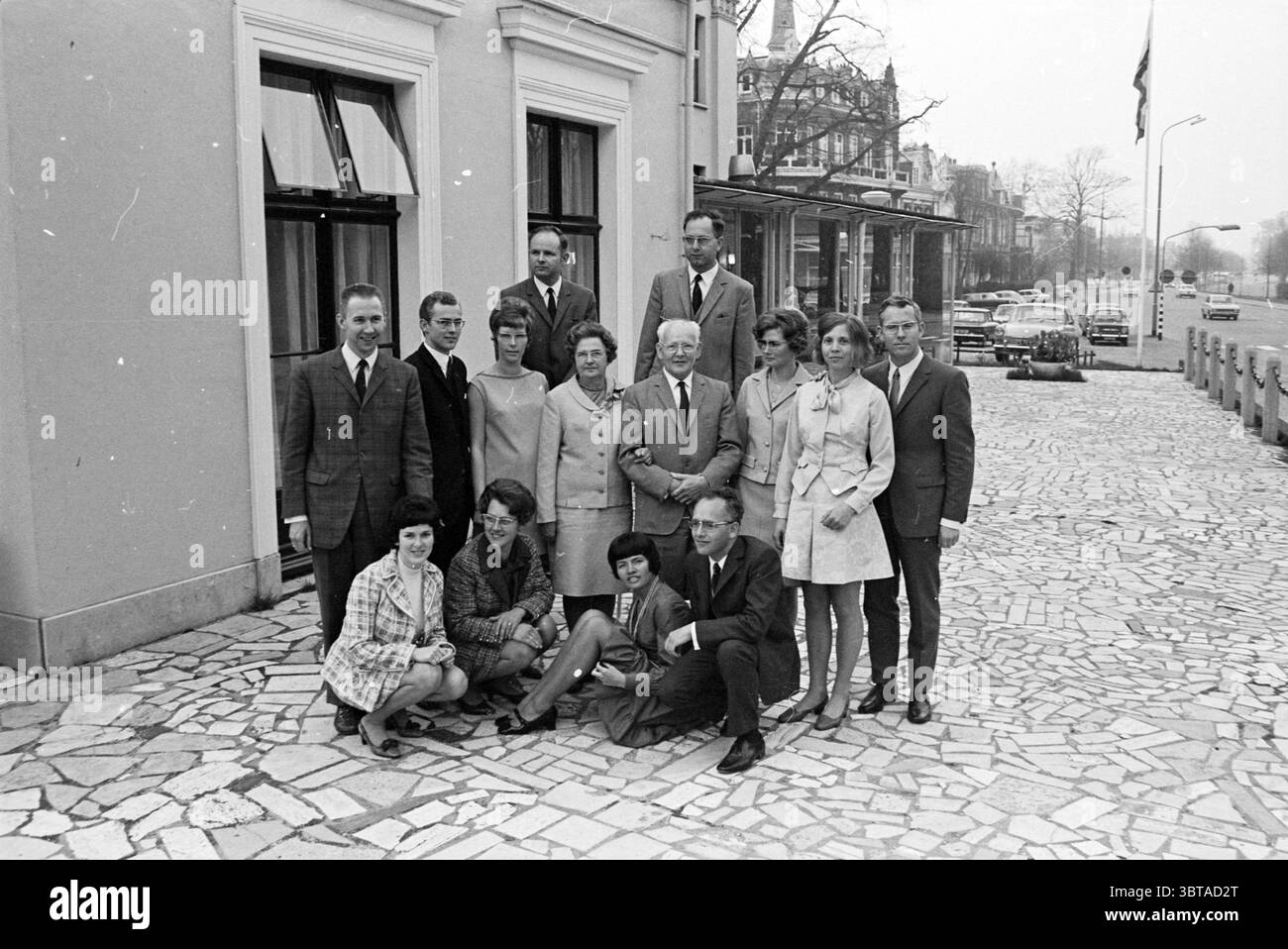 Photo de famille des familles Piet, Whizgle News, Dutch Desk, pays-Bas, 1950 - 2000 le 02-05-1969. Ces rubriques sont illustrées dans l'image. La scène capture un groupe de personnes posées à l'extérieur d'un bâtiment élégant avec un style architectural typique du design européen du milieu du XXe siècle. L'extérieur présente des murs en pierre de couleur claire et de grandes fenêtres, reflétant un sentiment de sophistication. Au premier plan, un groupe diversifié de quatorze individus se dresse en deux rangées. La première rangée se compose d'un mélange d'hommes et de femmes. Les femmes sont habillées dans une variété de tenues à la mode, avec s Banque D'Images
