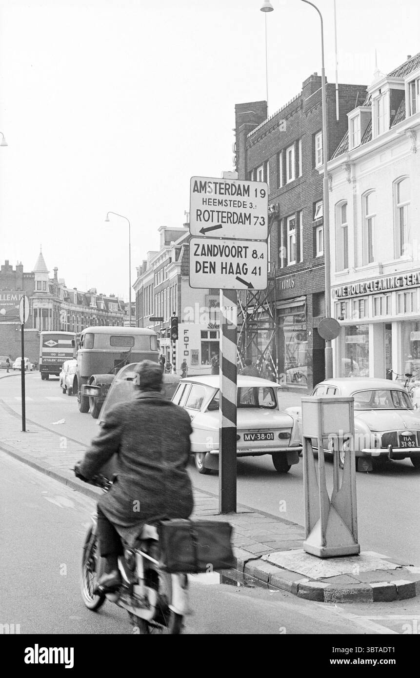 Panneau avec flèches Houtplein Traffic Safe Traffic Haarlem Houtplein pays-Bas, Whizgle News, Dutch Desk, pays-Bas, 1950 - 2000 le 17-03-1965. Ce sont les éléments de l'image. La scène représente une rue urbaine animée, caractérisée par un mélange d'architecture historique et du milieu du XXe siècle. Les bâtiments bordant la rue vont de façades en briques à des designs plus contemporains, avec un mélange de devantures de magasins et d'espaces résidentiels. Au premier plan, un cycliste vêtu d'un long manteau sombre navigue devant une rangée de voitures anciennes garées, leurs formes douces et arrondies indicatives de l'époque. Le bicyc Banque D'Images
