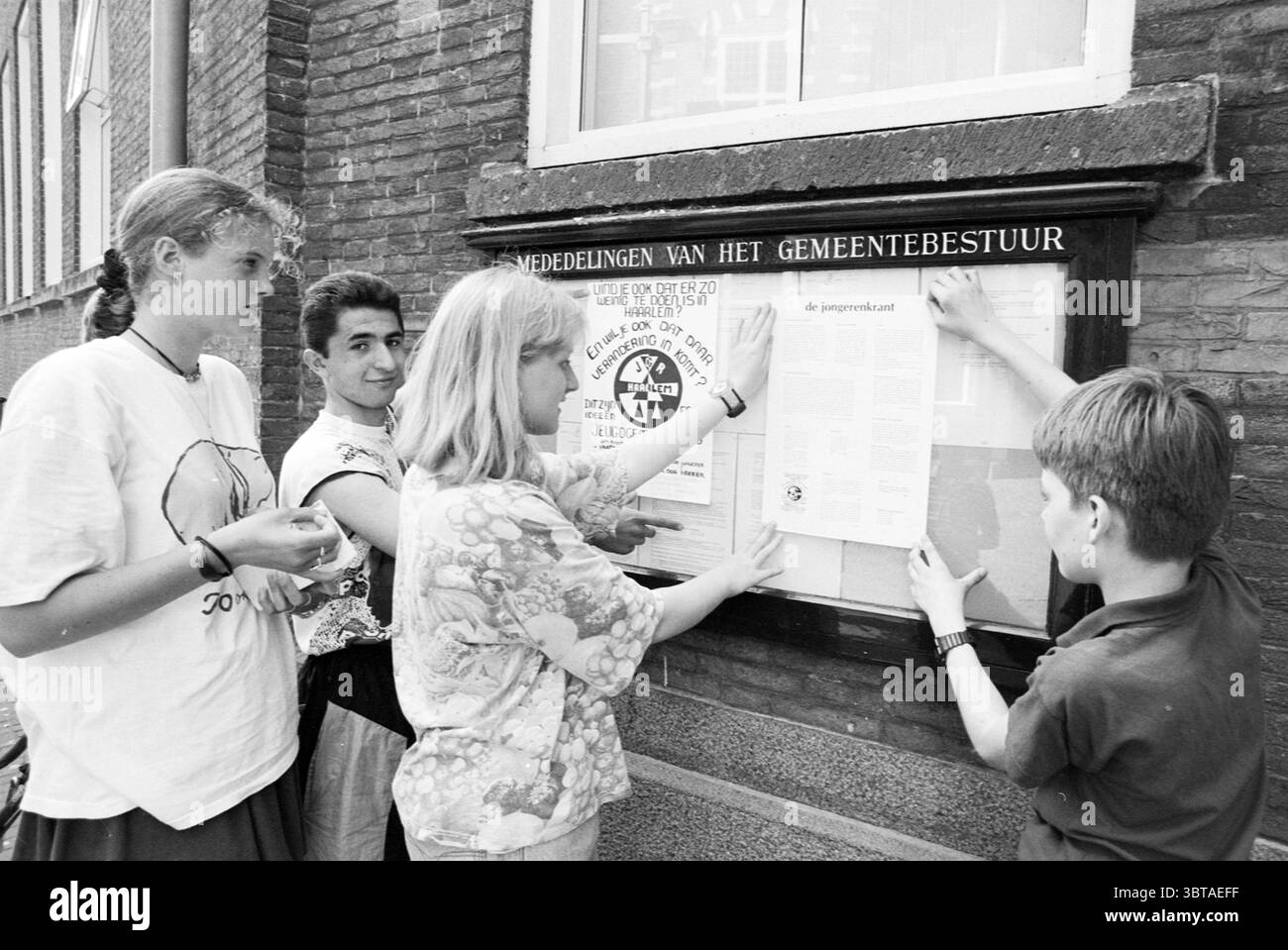 Journal jeunesse conseil municipal des jeunes, Whizgle News, Dutch Desk, pays-Bas, 1950 - 2000 le 06-06-1996. L'image inclut ces rubriques. Dans un cadre urbain animé, un groupe de jeunes s’engage activement autour d’un panneau d’affichage fixé à un mur de briques. Le mur est composé d'un mélange de briques brunes et rouges, montrant une certaine usure avec quelques fissures soulignant son âge. Le tableau d'affichage est en bois avec une façade en verre, contenant divers avis et prospectus, dont certains sont juste apposés par le groupe. Les individus sont un mélange d'adolescents, chacun portant des vêtements décontractés qui reflètent leur vous Banque D'Images