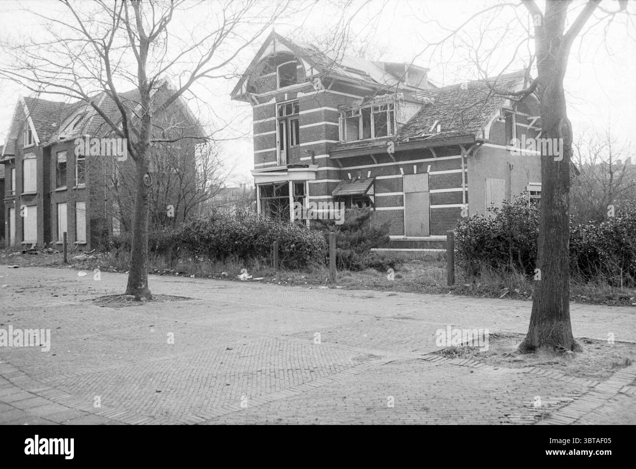 Villa Nooitgedagt Kanaalstraat IJmuiden maisons et construction de maisons IJmuiden Kanaalstraat pays-Bas, Whizgle News, Dutch Desk, pays-Bas, 1950 - 2000 on 19-01-1988. L'image inclut ces rubriques. La scène représente un environnement urbain calme et un peu désolé. Sur la gauche, un bâtiment de deux étages montre des signes de négligence, avec des fenêtres en bois et une façade altérée, présentant un mélange de briques et de plâtre. Le style du bâtiment a un charme classique, mais son état indique l'abandon. À côté de lui, une structure de style similaire se dresse, également vacante et en mauvais état, avec envahi ve Banque D'Images