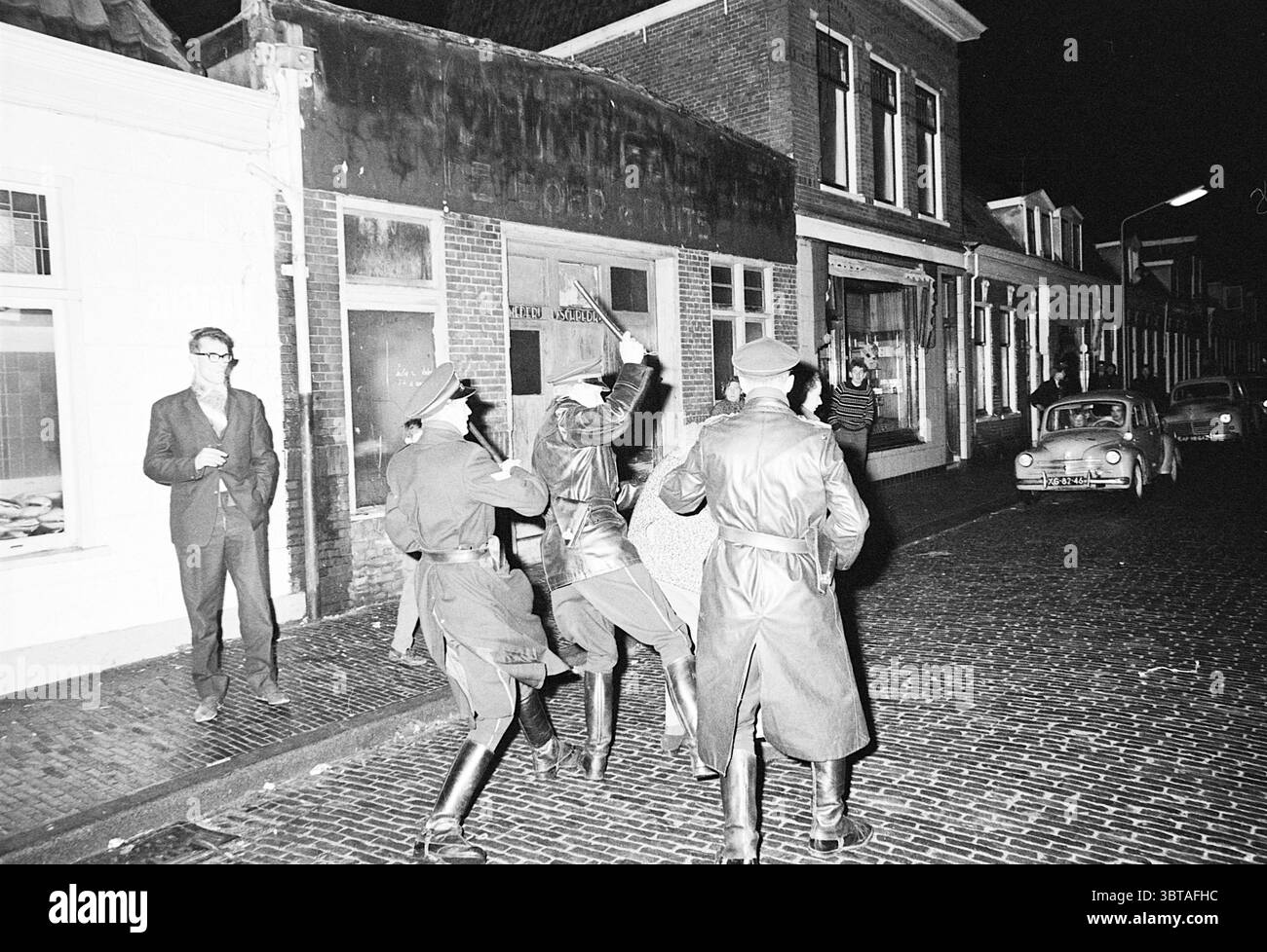 Arbres de Noël à Haarlem + les émeutes du réveillon du nouvel an les pompiers, Whizgle News, Dutch Desk, pays-Bas, 1950 - 2000 le 01-01-1965. Ces rubriques apparaissent dans l'image. Dans une rue urbaine faiblement éclairée, la scène capte un sentiment de tension et de signification historique. Les pavés brillent légèrement de l'éclat d'une pluie récente, reflétant la faible lueur des lampadaires projetant une lumière chaude et silencieuse. Trois personnages dans des tenues sombres de style militaire dominent le premier plan, leurs chapeaux baissés, créant un sentiment d'anonymat et d'autorité. Ils sont engagés dans une pose dynamique, peut-être impliqué moi Banque D'Images