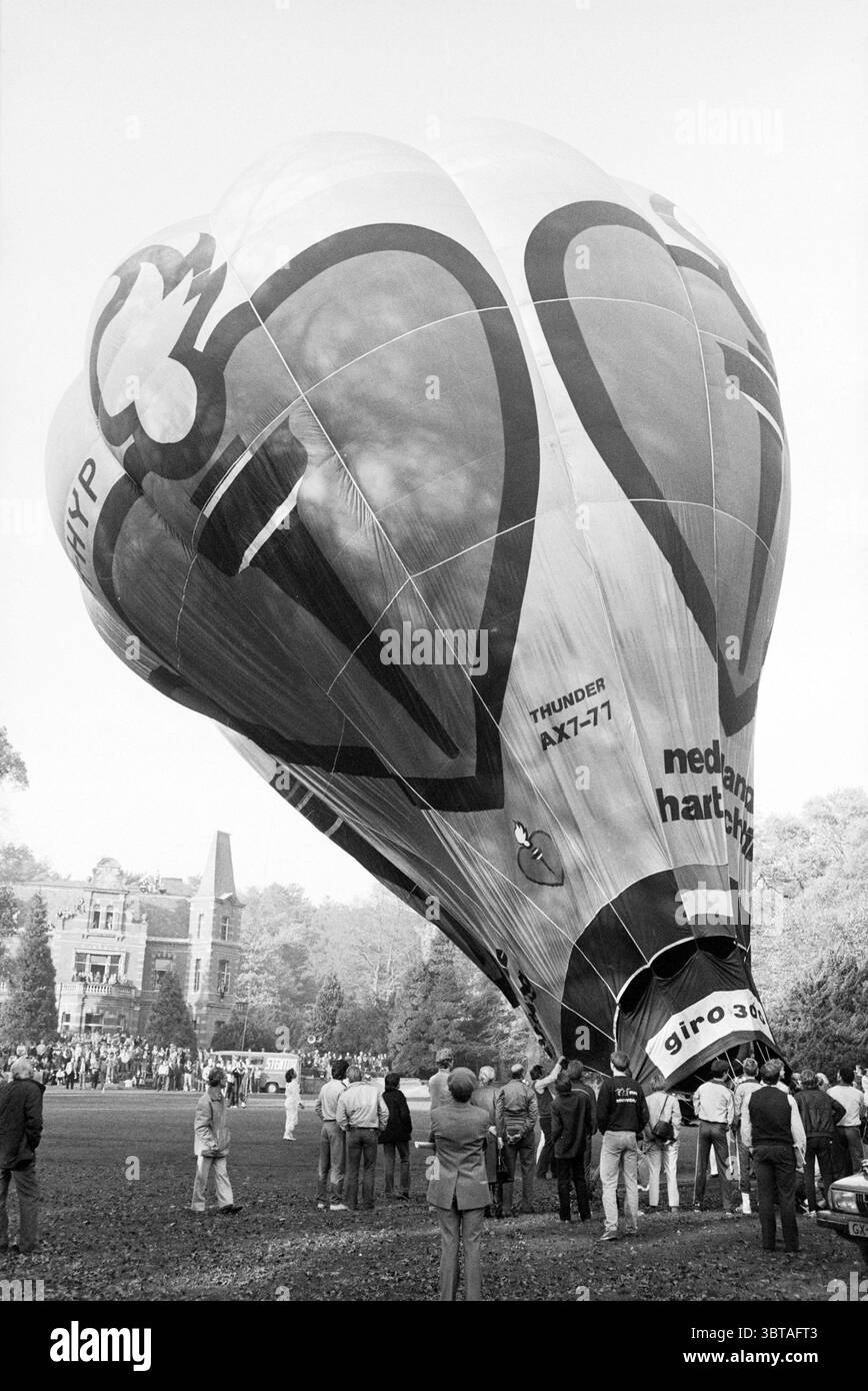 Secrétaire d'Etat v.d Reijden en ballon et MRS Ginjaar-Maas C.I.O.S., Whizgle News, Dutch Desk, pays-Bas, 1950 - 2000 on 10-01-1983. Voici les rubriques de l'image. La scène met en scène une montgolfière vibrante qui domine le premier plan, son tissu massif bougeant vers le haut. Le ballon présente des couleurs vives, avec un design joyeux incorporant des formes rouges et blanches, qui rappellent peut-être des coeurs ou des nuages. Sa grande taille lui confère une présence imposante sur toile de fond. En arrière-plan, un bâtiment majestueux à l'architecture complexe peut être vu, suggérant un historique ou e Banque D'Images