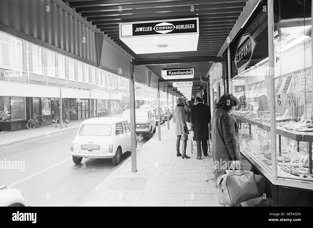 Vue d'ensemble de Cronjéstraat avec canopées Cronjestraat Haarlem Haarlem Generaal Cronjéstraat pays-Bas, Whizgle News, Dutch Desk, pays-Bas, 1950 - 2000 sur 12-12-1969. L'image contient ces rubriques. La scène représente une rue urbaine animée avec un accent sur une rangée de magasins sous une passerelle couverte. L'architecture présente un auvent dans un ton sourd, projetant de douces ombres le long du trottoir. Sur le côté gauche, divers affichages présentent un éventail de marchandises, scintillant sous un éclairage vif. Les objets décoratifs et les bijoux sont méticuleusement disposés, mettant en valeur leurs conceptions complexes. Le g Banque D'Images