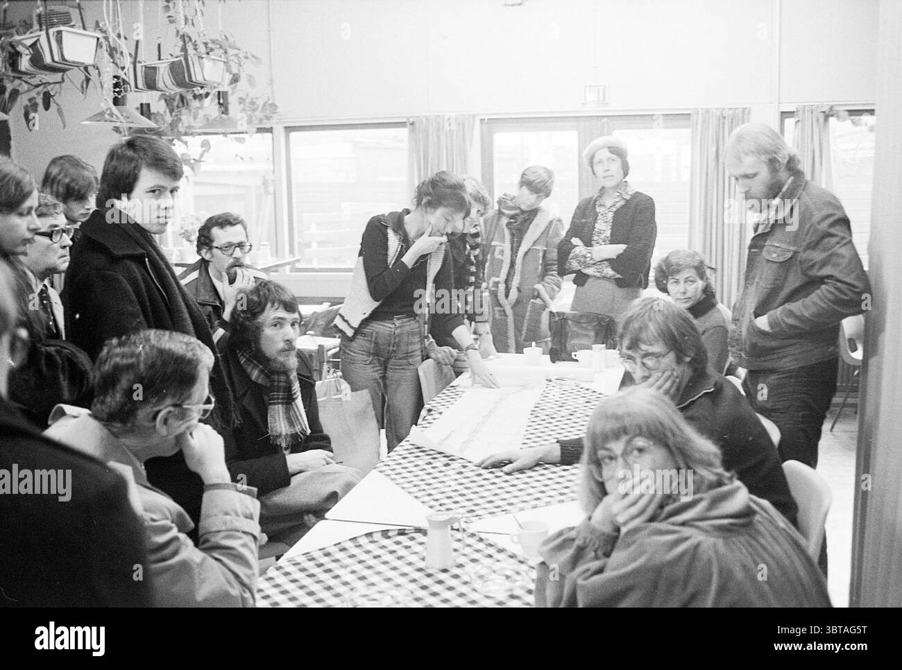 Excursion Leidseplein excursion, Whizgle News, Dutch Desk, pays-Bas, 1950 - 2000 on 30-11-1977. Ces rubriques sont illustrées dans l'image. La scène capture un rassemblement dans une pièce spacieuse et très éclairée, avec de grandes fenêtres permettant à la lumière naturelle de filtrer. L’atmosphère est vivante mais contemplative, avec un air de collaboration. Au centre, une longue table est ornée d’une nappe à carreaux en noir et blanc, servant de point focal à l’assemblage. Plusieurs individus sont assis autour de la table, certains penchant en avant par intérêt, tandis que d'autres sont profondément dans la pensée. Il y a une gamme o Banque D'Images
