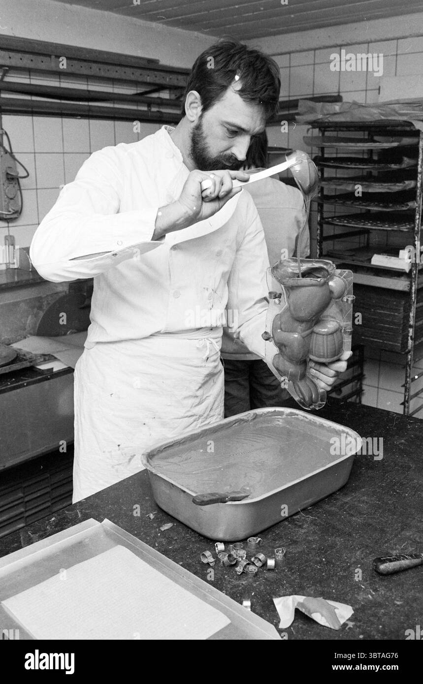 Fabrication de chocolat lapin de Pâques fa. Hazenberg IJmuiden Palm Pâques et coutumes de Pâques IJmuiden pays-Bas, Whizgle News, Dutch Desk, pays-Bas, 1950 - 2000 on 28-03-1986. Voici les rubriques de l'image. La scène représente un homme dans un environnement de cuisine professionnel, immergé dans une tâche culinaire. Il a une barbe soigneusement soignée et est habillé d'un manteau de chef blanc, qui est légèrement taché, indiquant une éthique de travail pratique. La cuisine a une atmosphère propre mais animée, avec des comptoirs en acier inoxydable et des étagères doublées de divers plateaux de cuisson et ustensiles. Dans ses mains, il tient un un Banque D'Images