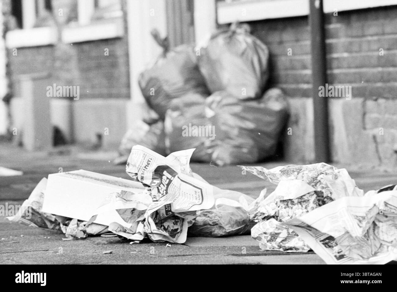Ordures à côté des sacs poubelle Parklaan Haarlem Parklaan pays-Bas, Whizgle News, Dutch Desk, pays-Bas, 1950 - 2000 on 06-12-1979. L'image inclut ces rubriques. La scène présente un environnement urbain graveleux, caractérisé par un trottoir encombré de journaux jetés et de boîtes en carton. Ces matériaux sont froissés et dispersés de manière désordonnée, suggérant un usage récent ou une élimination rapide. Les journaux, avec leurs images monochromes emblématiques, font allusion à des histoires ou publicités précédentes, tandis que les boîtes en carton semblent usées et grises. En arrière-plan, plusieurs grands sacs poubelles, un Banque D'Images