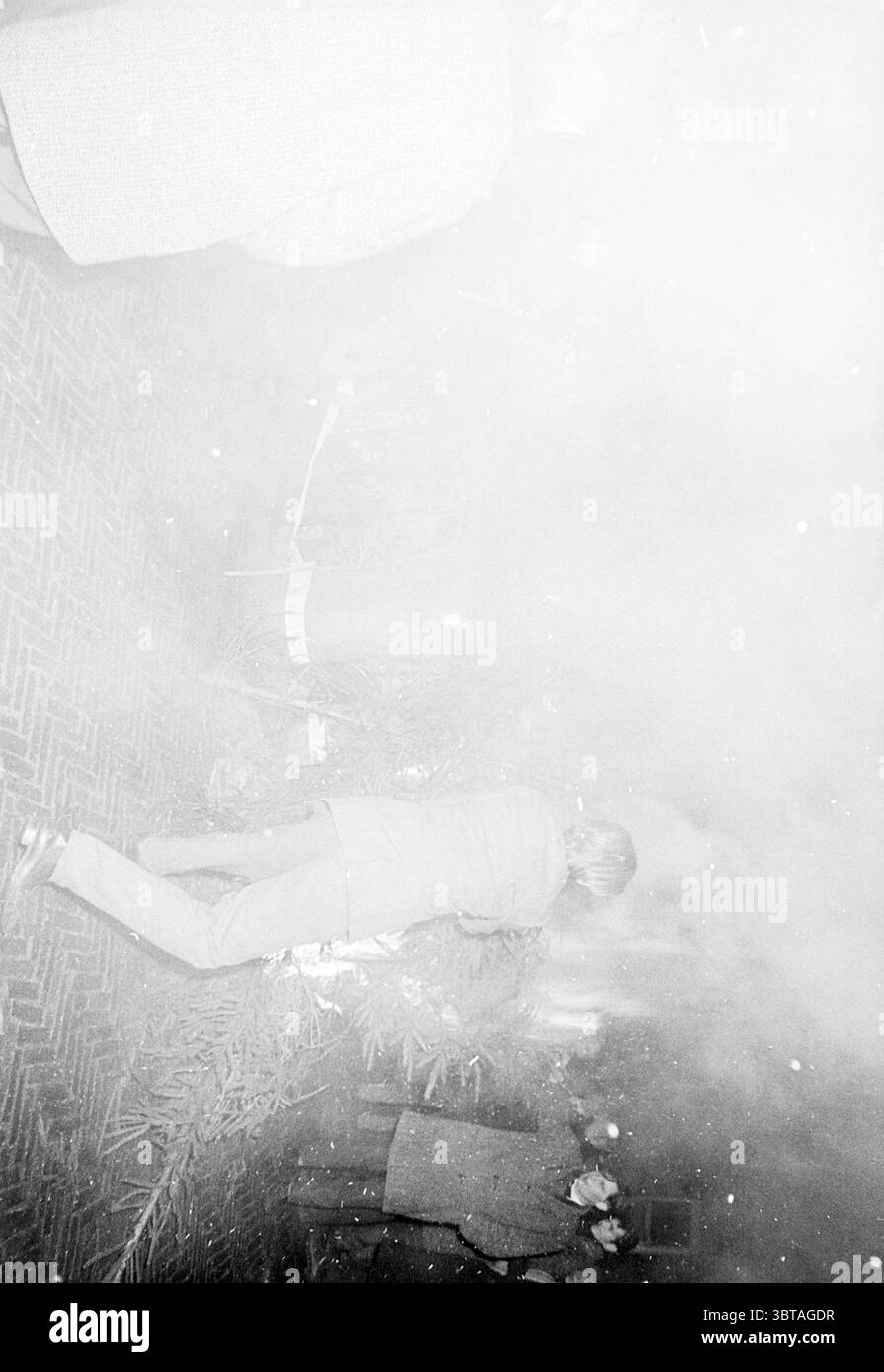 Arbres de Noël à Haarlem + les émeutes du réveillon du nouvel an les pompiers, Whizgle News, Dutch Desk, pays-Bas, 1950 - 2000 le 01-01-1965. Ces rubriques apparaissent dans l'image. La scène capture un moment brumeux et atmosphérique, probablement placé à l'intérieur avec une mise au point douce qui lui donne une qualité éthérée. L'éclairage est faible, projetant une douce lueur qui rehausse l'ambiance brumeuse. Au premier plan, une personne vêtue d'une tenue de couleur claire peut être vue se pencher, le dos tourné vers le spectateur. Leur posture suggère qu'ils sont engagés dans une activité, éventuellement arranger ou observer quelque chose sur le gr Banque D'Images