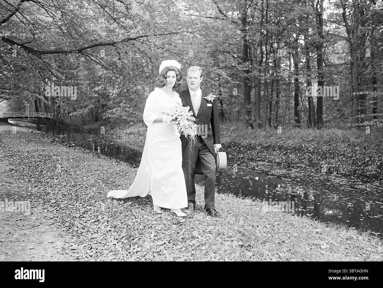 Mariage v.d. lit - Hendriks mariage, Whizgle News, Dutch Desk, pays-Bas, 1950 - 2000 on 11-10-1968. Voici les rubriques de l'image. Dans un cadre extérieur serein, un couple se tient ensemble au bord d'un ruisseau qui coule doucement. La femme porte une élégante robe de mariée à manches longues qui coule gracieusement au sol, complétée par un chapeau frappant et élégant orné d'un large bord. Son bouquet, rempli de feuillage luxuriant et de fleurs délicates, est bercé dans ses bras, ajoutant une touche de dynamisme à son ensemble. À côté d'elle, l'homme est habillé d'un costume noir classique, avec un noeud t Banque D'Images