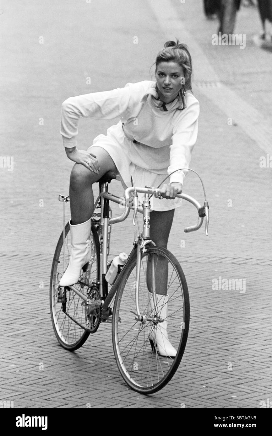 Tendances mode, Whizgle News, Dutch Desk, pays-Bas, 1950 - 2000 sur 04-09-1982. Ces rubriques apparaissent dans l'image. Une jeune femme est capturée à mi-mouvement sur un vélo, exsudant la confiance et un sentiment de spontanéité ludique. Elle est habillée dans une tenue élégante et entièrement blanche qui se compose d'un haut ajusté et d'une jupe évasée, créant un contraste marqué avec ses bottes blanches saisissantes hauteur genou. L'ensemble met en valeur sa sensibilité avant-gardiste. Le vélo lui-même a un design classique, avec un cadre léger qui est typique de la conduite urbaine. Les roues sont étroites, et le guidon Banque D'Images