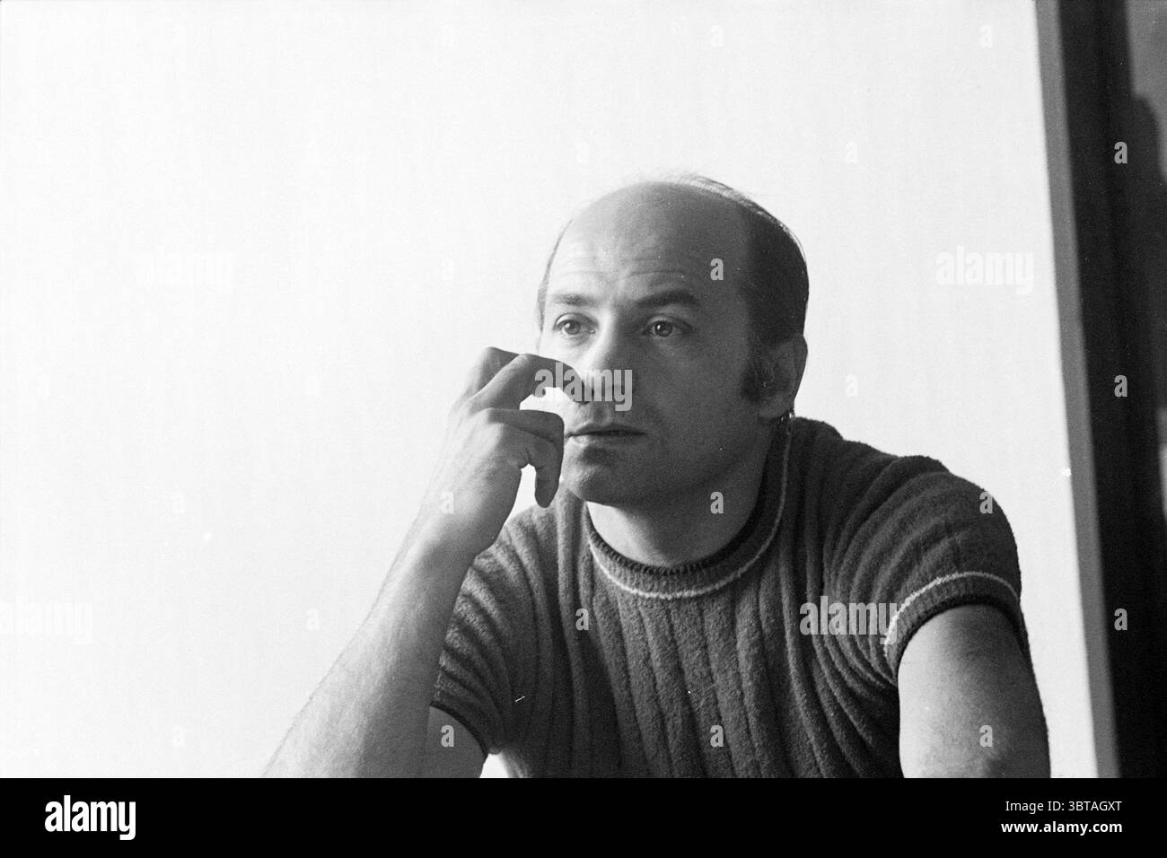 Photo d'un homme., Whizgle News, Dutch Desk, pays-Bas, 1950 - 2000. L'image contient ces rubriques. La scène montre un homme assis avec soin, son corps tourné légèrement à l'écart de la source de lumière. Il a un visage arrondi, une ligne de cheveux reculée et une expression contemplative, indiquant qu'il est profondément dans la pensée. Sa main repose sur son visage, un doigt touchant doucement ses lèvres, suggérant une introspection. Il est habillé d'un pull en maille texturée ajusté qui semble être une nuance discrète, peut-être grise ou marine, améliorant l'ambiance décontractée mais réfléchie de la scène. L'environnement environnant Banque D'Images