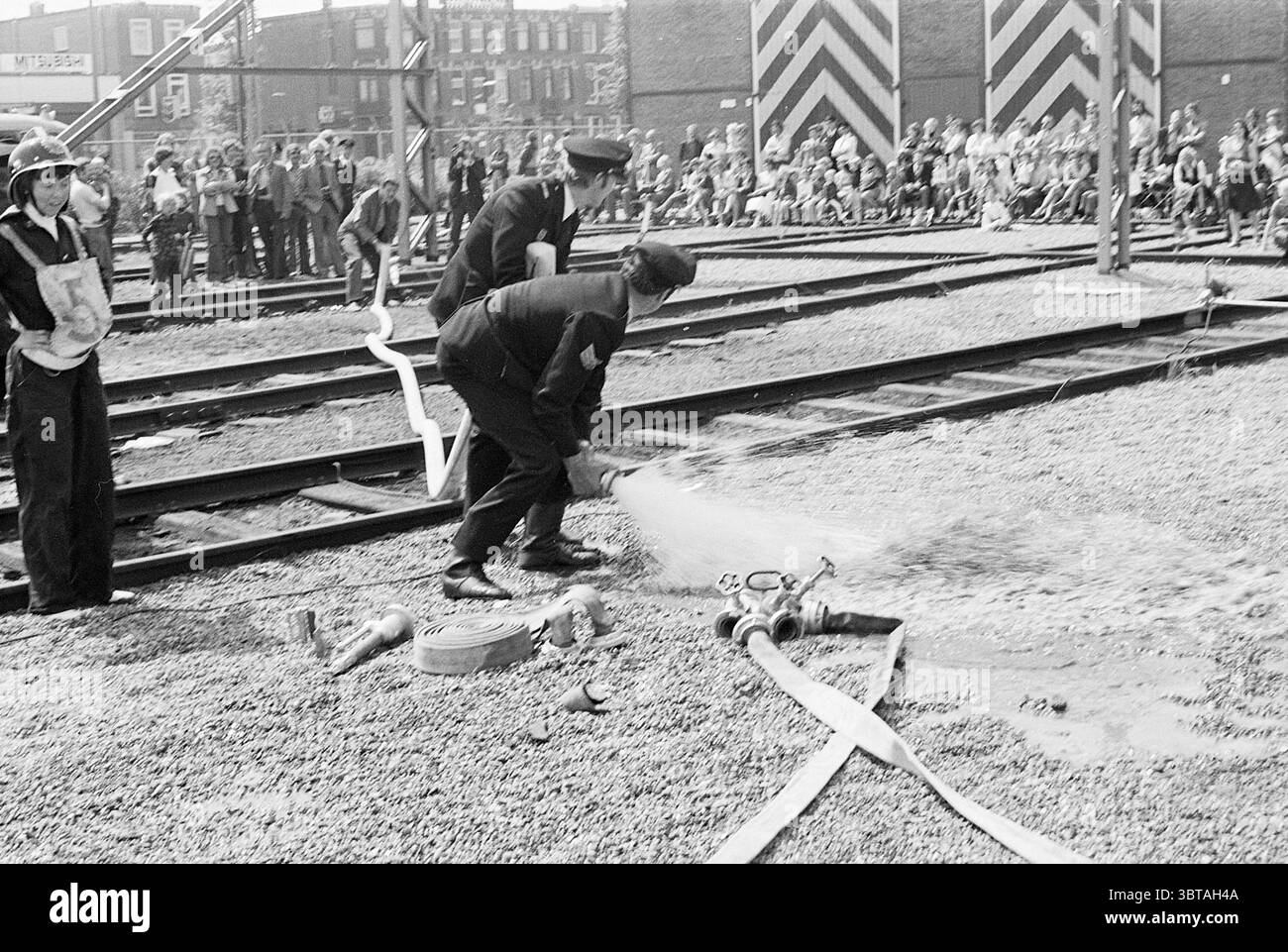 Compétitions de pompiers équipe féminine Haarlem Fires Fire brigade Competitions, Whizgle News, Dutch Desk, pays-Bas, 1950 - 2000 le 15-05-1976. L'image contient ces rubriques. Dans un cadre extérieur animé, une foule s'est rassemblée le long du bord d'une gare de triage. La scène est dominée par le contraste saisissant de la lumière et de l'ombre, suggérant une journée lumineuse remplie d'activité. Deux personnages, vêtus d'uniformes sombres indiquant l'autorité ou le service d'urgence, participent activement à une manifestation de lutte contre l'incendie. Ils sont positionnés au premier plan, axés sur un grand tuyau d'incendie enroulé fr Banque D'Images
