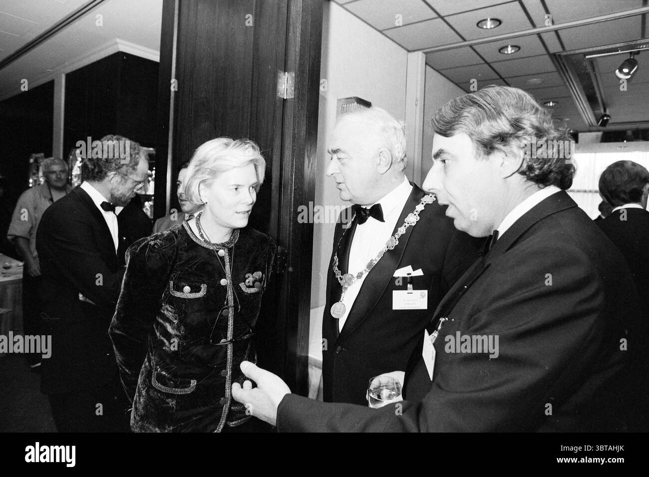 Réunion de la Chambre de commerce à Schiphol Schiphol, Whizgle News, Dutch Desk, pays-Bas, 1950 - 2000 le 19-09-1991. Ce sont les éléments de l'image. La scène se déroule dans un cadre intérieur élégant, caractérisé par une ambiance sophistiquée et une opulence subtile. Les murs sont ornés de lambris sombres, créant une atmosphère chaleureuse mais formelle, tandis que l'éclairage doux des luminaires suspendus projette une douce lueur dans la pièce. Au premier plan, trois personnes engagent la conversation. Une femme, vêtue d'une tenue chic et sombre avec une texture de velours, se tient les bras doucement croisés, portant un Banque D'Images