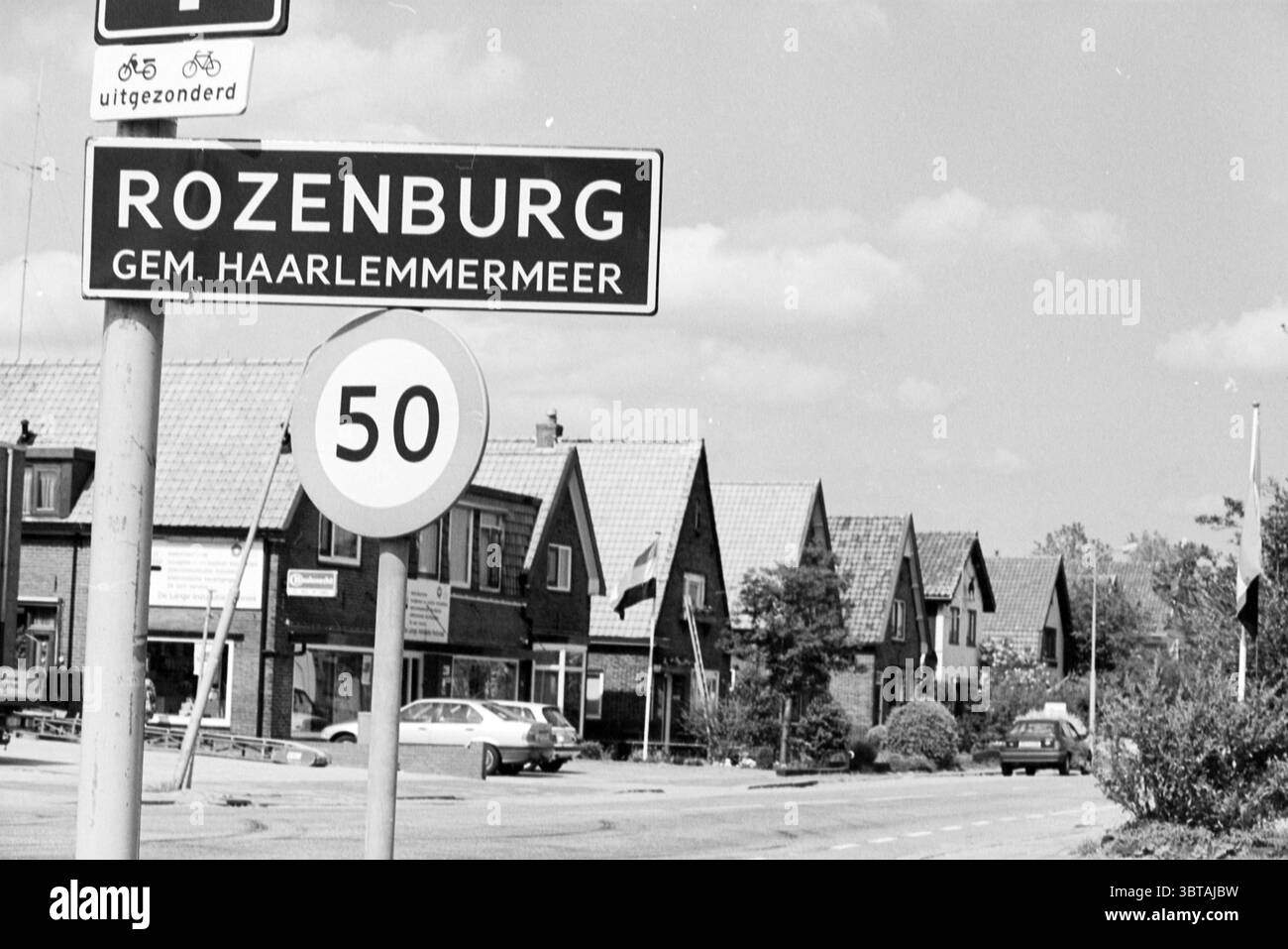 Village Rozenburg Rozenburg, Whizgle News, Dutch Desk, pays-Bas, 1950 - 2000 on 04-05-1993. Ces rubriques apparaissent dans l'image. La scène capture une rue calme dans une zone de banlieue, bordée de maisons modestes affichant des toits angulaires et des façades à pignons. L'architecture présente un mélange de styles, avec certaines maisons ornées de petits jardins, tandis que d'autres présentent des allées pavées. Au-dessus, le ciel est une douce nuance de gris, faisant allusion au temps couvert, ce qui ajoute une atmosphère tamisée au cadre. Une route traverse le premier plan, où les panneaux de signalisation sont bien en vue. Un panneau indique une vitesse Banque D'Images