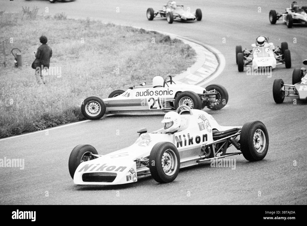 Course automobile sur Zandvoort., Whizgle News, Dutch Desk, pays-Bas, 1950 - 2000. L'image inclut ces rubriques. La scène capture un moment dynamique d'un événement de sport automobile sur un circuit sinueux. Plusieurs petites voitures de course élégantes, peintes dans différentes couleurs vives, zooment le long de la courbe de la piste, mettant en valeur leurs conceptions aérodynamiques. Au premier plan, une voiture marquée d'un logo remarquable court devant, sa peinture blanche brillante sur fond de tons discrets. Le pilote porte un casque reflétant la concentration intense et la compétition du moment. A proximité, une autre voiture dans une teinte de bleu profond est sli Banque D'Images