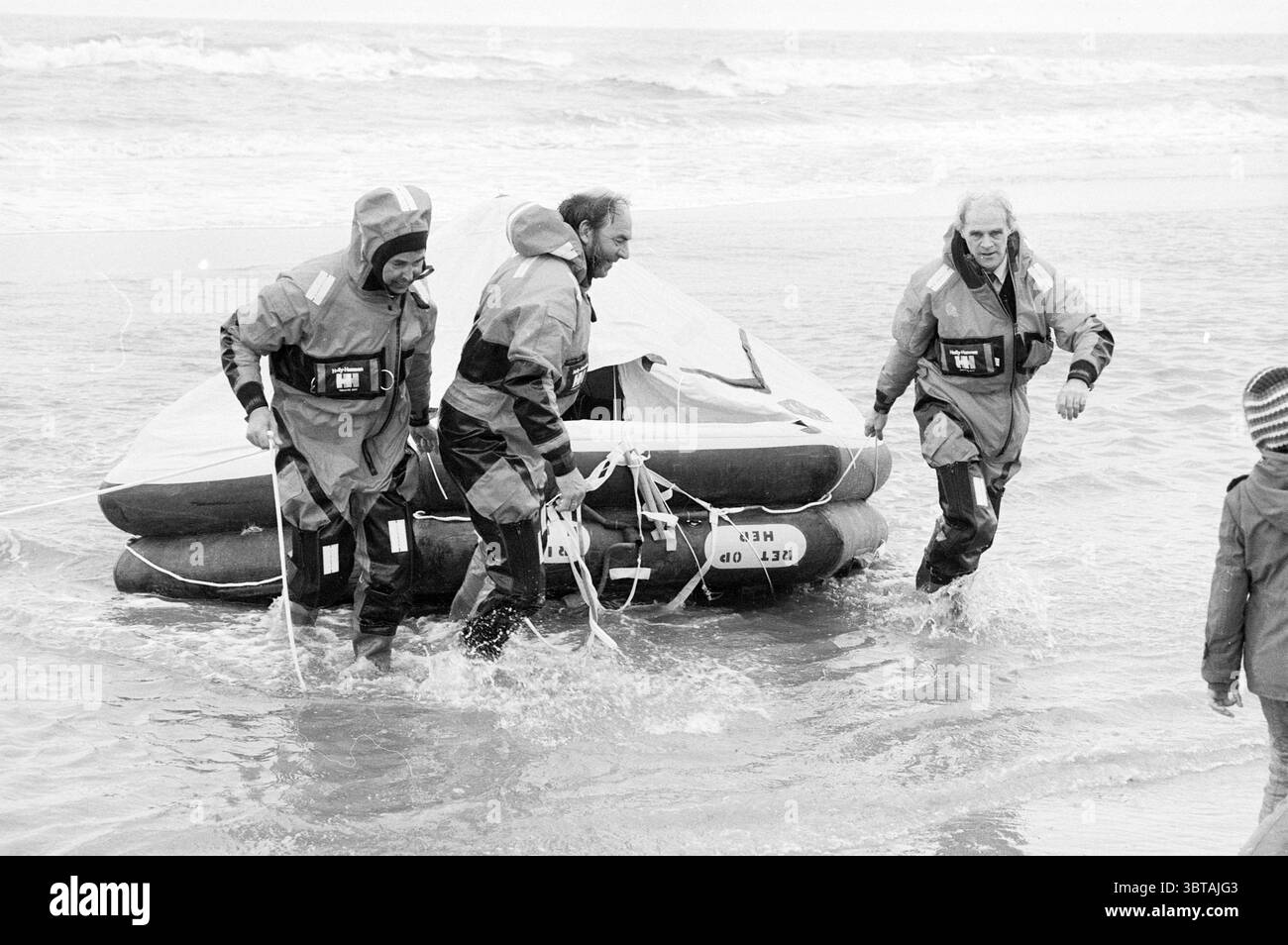 Exercice de sauvetage plage Zandvoort en relation avec TV médecine Rescue brigade service de sauvetage télévision Zandvoort, Whizgle News, Dutch Desk, pays-Bas, 1950 - 2000 on 11-04-1983. L'image montre ces rubriques. La scène capture un moment dynamique sur une plage, où trois individus émergent de l’océan. Tous sont revêtus de costumes sombres et imperméables qui brillent avec l'humidité de la mer. Chaque personne semble concentrée et déterminée, naviguant dans des eaux peu profondes qui éclaboussent autour de leurs jambes. Au centre, deux des individus manœuvrent un petit radeau gonflable, qui est Banque D'Images