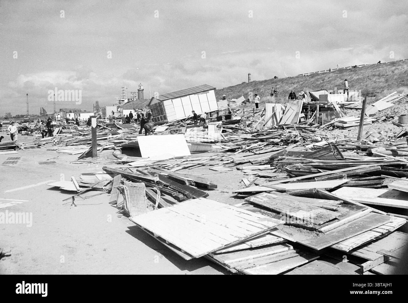 Déblayer les gravats des maisons d'été sur la plage d'IJmuiden tempête et dégâts causés par la tempête plage et plages IJmuiden Nederland, Whizgle News, Dutch Desk, pays-Bas, 1950 - 2000 on 21-04-1980. Ces rubriques sont illustrées dans l'image. La scène capture un paysage marqué par la dévastation, où des débris et des restes de structures se trouvent dispersés sur un terrain sablonneux. Des piles de planches de bois, de panneaux et de matériaux de construction brisés sont éparpillés de manière chaotique, formant un arrangement presque abstrait qui suggère une calamité récente. Le sol est largement atténué, comprenant diverses nuances de brun et de beige du Banque D'Images