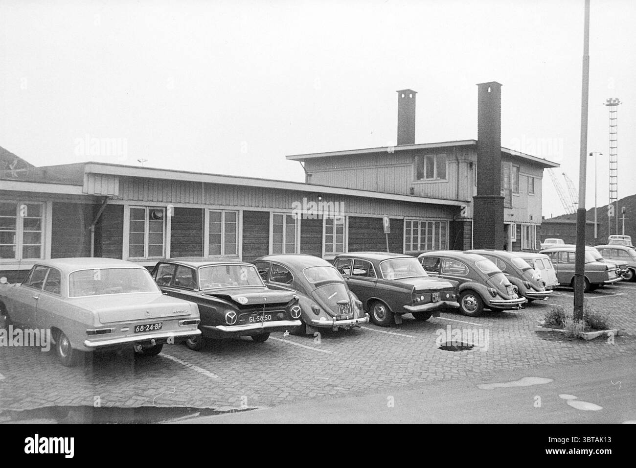Construction des hauts fourneaux., Whizgle News, Dutch Desk, pays-Bas, 1950 - 2000. L'image contient ces rubriques. La scène montre une ligne de voitures anciennes garées sur un terrain pavé devant un modeste bâtiment de deux étages. L'architecture de la structure présente un design simple avec une combinaison de grandes fenêtres et de panneaux de bois, évoquant un style du milieu du XXe siècle. Le bâtiment a deux cheminées sombres s'élevant de son toit, ajoutant une touche rustique. Les voitures, pour la plupart des modèles classiques, affichent une gamme de couleurs discrètes — gris, blancs et tons pastel — évoquant une atmosphère nostalgique. Certains Banque D'Images