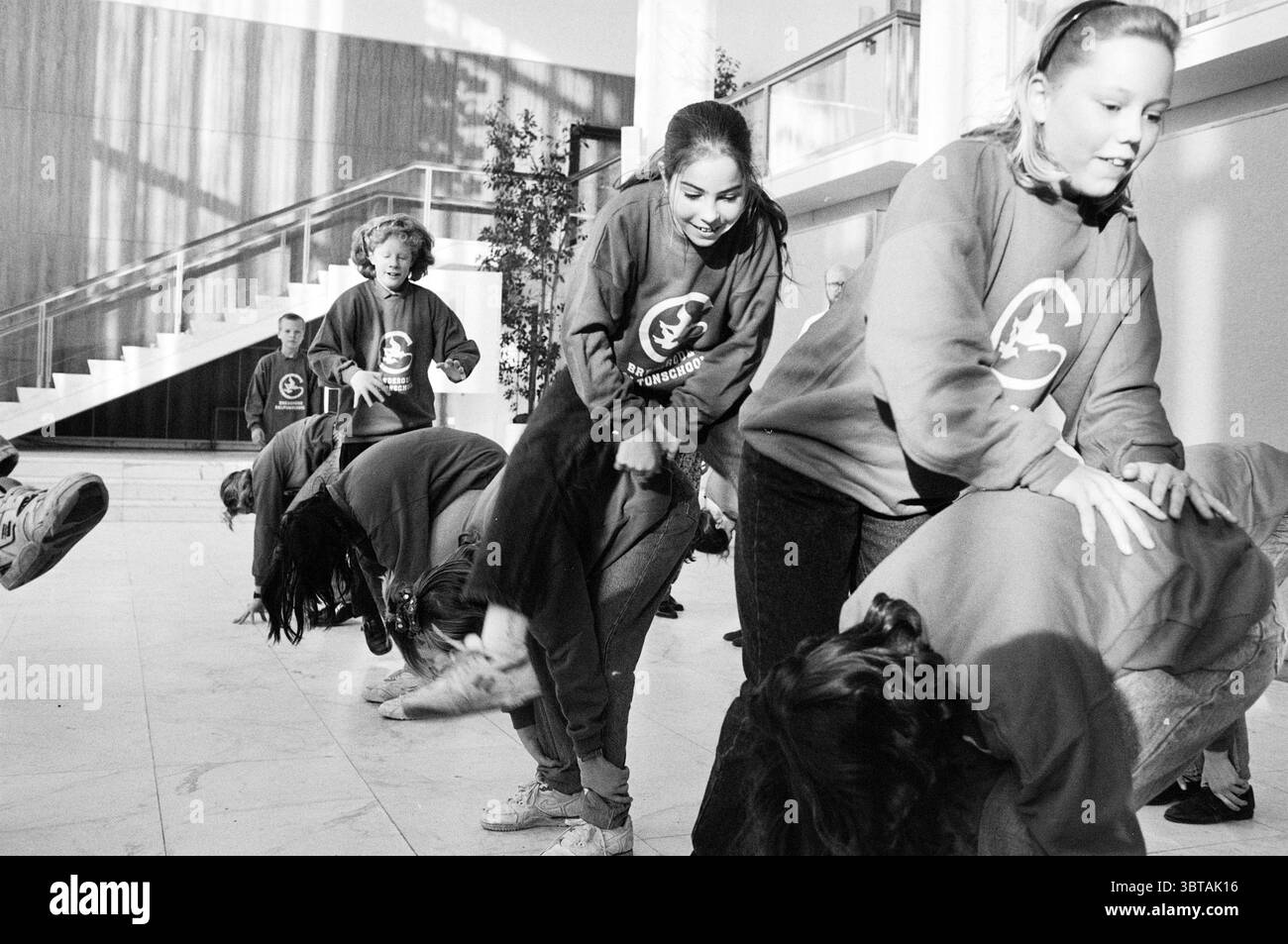 Classe de gym Brederodeschool IJm mairie. IJmuiden pays-Bas, Whizgle News, Dutch Desk, pays-Bas, 1950 - 2000 on 29-11-1991. L'image contient ces rubriques. Dans un cadre intérieur spacieux caractérisé par une architecture moderne, un groupe d'enfants est engagé dans une activité ludique, créant une atmosphère vivante et dynamique. La grande zone présente de hauts plafonds et une grande lumière naturelle qui pénètre à travers de hautes fenêtres, projetant des ombres douces sur le sol poli. Les enfants, vêtus de sweat-shirts surdimensionnés assortis dans une nuance de rouge vif, sont à différentes étapes d'un jeu de groupe énergique Banque D'Images