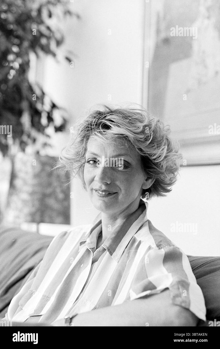 Portrait Liedwien Rolink Santpoort Santpoort, Whizgle News, Dutch Desk, pays-Bas, 1950 - 2000 le 14-05-1992. Ce sont les éléments de l'image. La scène met en scène une femme assise confortablement sur un canapé moelleux, dégageant un sentiment de calme et de convivialité. Ses cheveux sont coiffés de boucles volumineuses et de couleur claire qui encadrent magnifiquement son visage, attirant l'attention sur son sourire doux et ses yeux expressifs. Elle porte un chemisier rayé dans des tons pastel doux, qui ajoute une sensation de légèreté et de luminosité à la composition globale. L'environnement environnant est légèrement éclairé, suggérant un chaleureux, invitant a Banque D'Images