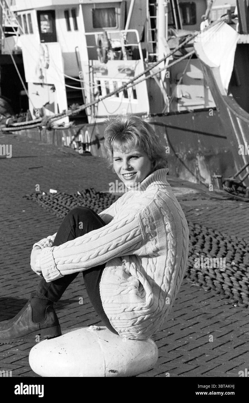 Marianne Verschuuren port of IJmuiden IJmuiden pays-Bas, Whizgle News, Dutch Desk, pays-Bas, 1950 - 2000 on 04-12-1997. Ces rubriques apparaissent dans l'image. Dans un cadre de marina animé, une jeune femme est assise sur une grande pierre blanche arrondie, sa posture détendue et décontractée. Elle porte un pull en maille torsadée confortable et surdimensionné dans des tons clairs, qui contraste avec les teintes plus sombres de son pantalon ajusté. Ses cheveux courts et touffus ont une qualité douce et ludique, encadrant son visage alors qu'elle sourit de manière engageante envers le spectateur. La toile de fond présente un groupe de bateaux de pêche, aux couleurs vives, avec des teintes Banque D'Images