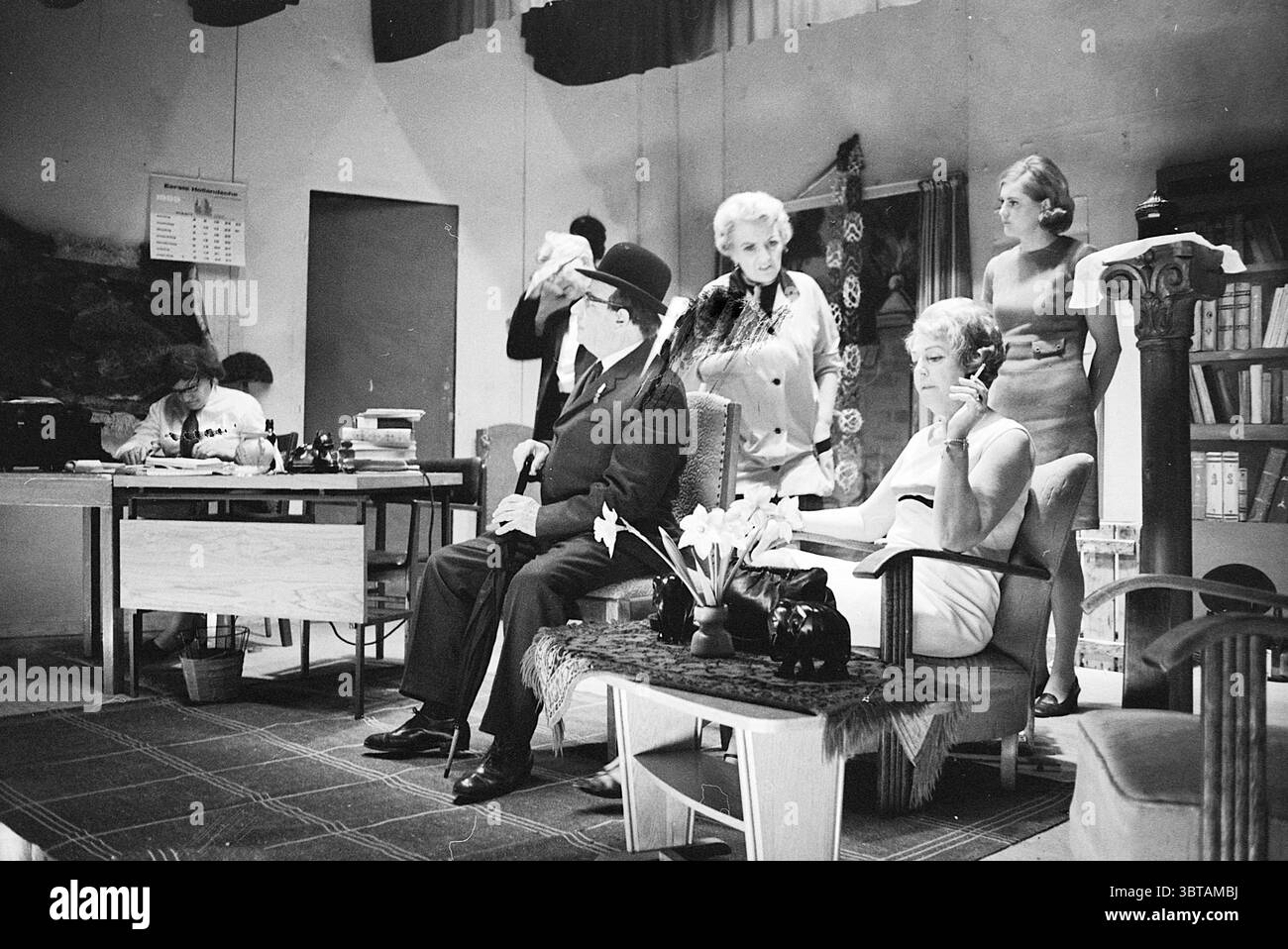 Représentation scénique., Whizgle News, Dutch Desk, pays-Bas, 1950 - 2000. L'image contient ces rubriques. La scène se déroule dans un intérieur faiblement éclairé, rappelant une scène de théâtre vintage. Une palette monochrome saisissante contribue à une atmosphère nostalgique. Au premier plan, une femme s'assoit élégamment sur une chaise en peluche, vêtue d'une robe élégante et ajustée. Sa coiffure est emblématique d'une époque révolue, avec des vagues douces qui encadrent son visage. Elle apparaît contemplative, peut-être engloutie dans la pensée en regardant vers le bas un petit vase de fleurs sur une table voisine, ce qui ajoute une touche délicate à l'ot Banque D'Images