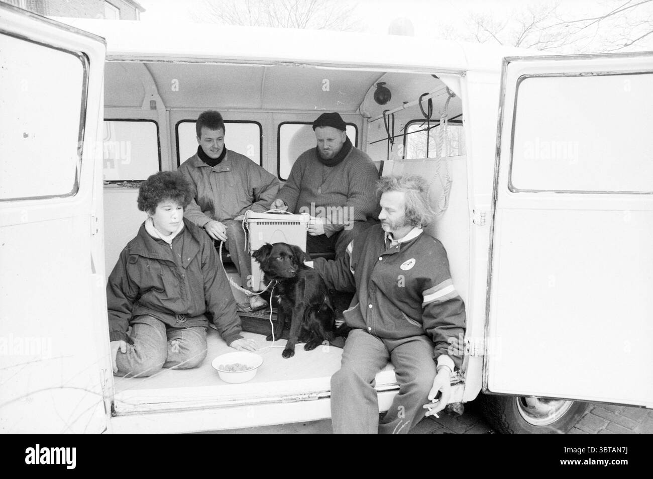 Animal Ambulance Schalkwijk Animals, Whizgle News, Dutch Desk, pays-Bas, 1950 - 2000 on 04-03-1987. L'image inclut ces rubriques. La scène représente un groupe de quatre individus assis à l'arrière d'un fourgon vintage, créant une atmosphère chaleureuse et intime. L’intérieur de la camionnette se caractérise par son design utilitaire, avec des murs métalliques et un sol simple, suggérant une utilisation pratique plutôt que le confort. Au premier plan, deux hommes sont assis de chaque côté d'un labrador noir, qui est concentré sur un bol de nourriture posé sur le sol. Le pelage brillant du chien contraste avec la douceur du véhicule Banque D'Images