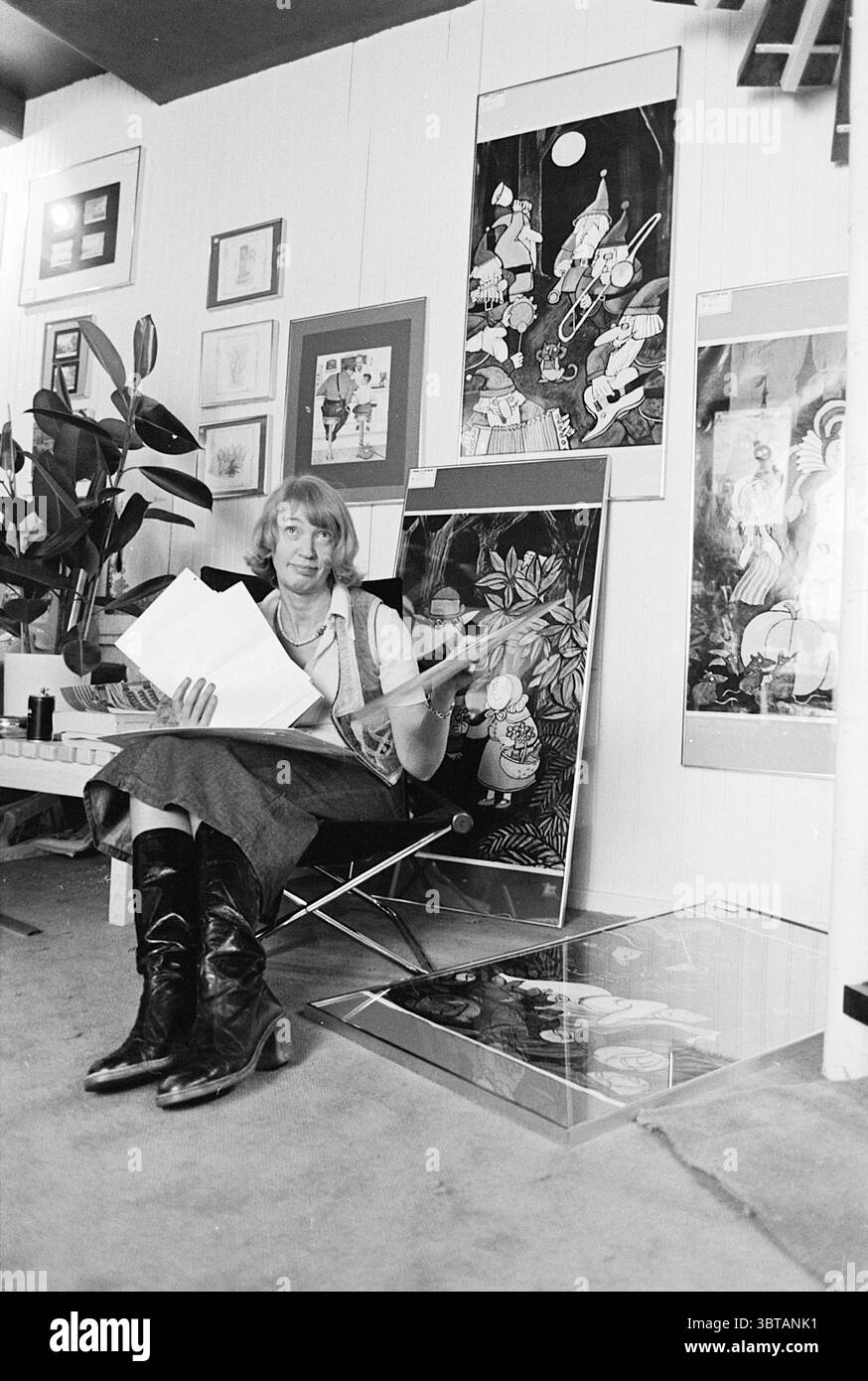 Shop 'Sign on the Wall' Amsterdam boutiques et kiosques, Whizgle News, Dutch Desk, pays-Bas, 1950 - 2000 sur 16-11-1976. Ce sont les éléments de l'image. Dans un studio d'art confortable, une femme s'assoit confortablement dans une chaise pliante, entourée d'un éventail d'œuvres d'art vibrantes. Elle a une apparence décontractée mais élégante avec un haut ajusté et une jupe longue qui coule jusqu'à son mi-mollet, complétée par des bottes noires brillantes hauteur genou qui attirent le regard. Son expression est réfléchie car elle tient une pile de papiers, probablement des esquisses ou des plans artistiques, les regardant attentivement, suggérant un moment de profonde concentratio Banque D'Images