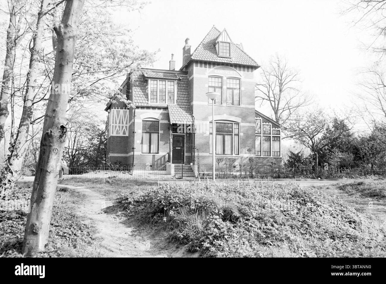 Ext. Wustelaan 90 Santpoort extérieur Santpoort Wüstelaan, Whizgle News, Dutch Desk, pays-Bas, 1950 - 2000 le 06-04-1990. Ce sont les éléments de l'image. La scène présente une charmante maison de trois étages caractérisée par ses caractéristiques architecturales uniques. Sa façade est un brun rougeâtre chaud, sur fond de verdure luxuriante. La maison affiche un mélange de toits à pignons abrupts, de conceptions complexes de fenêtres et d'éléments décoratifs, lui donnant un caractère vintage distinctif. Les environs sont invitants, avec un chemin de terre sinueux menant à l'entrée principale, flanqué de patc dense Banque D'Images
