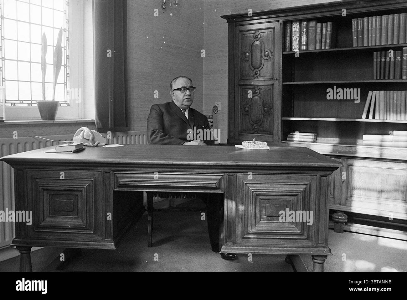 Portrait du nouveau maire Haarlemmerliede maires, Whizgle News, Dutch Desk, pays-Bas, 1950 - 2000 le 05-06-1968. Ces rubriques apparaissent dans l'image. Dans une chambre spacieuse, un homme est assis à un grand bureau en bois poli qui présente des sculptures complexes et un ton riche et chaleureux. Le bureau est soigneusement agencé avec un téléphone blanc et un petit objet décoratif, éventuellement un cendrier. Derrière lui, une grande étagère en bois est remplie de livres soigneusement empilés, certains avec des épines foncées et d'autres affichant des nuances plus claires, créant un contraste avec le bois plus sombre de l'étagère. Les murs sont ornés d'un doux, Banque D'Images